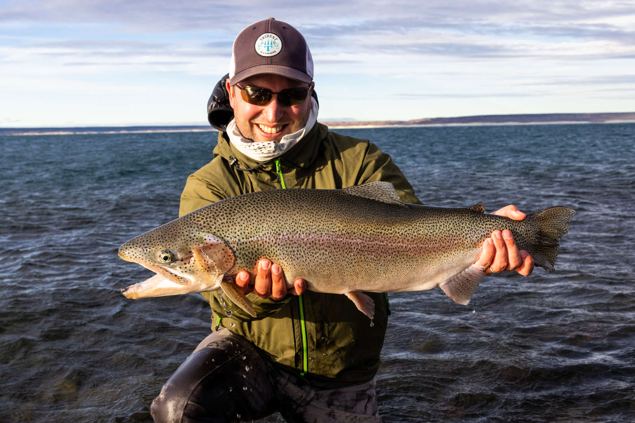 Smiling angler in olive green jacket and Trident cap holding large speckled trout at rocky lake shore, fishing trip