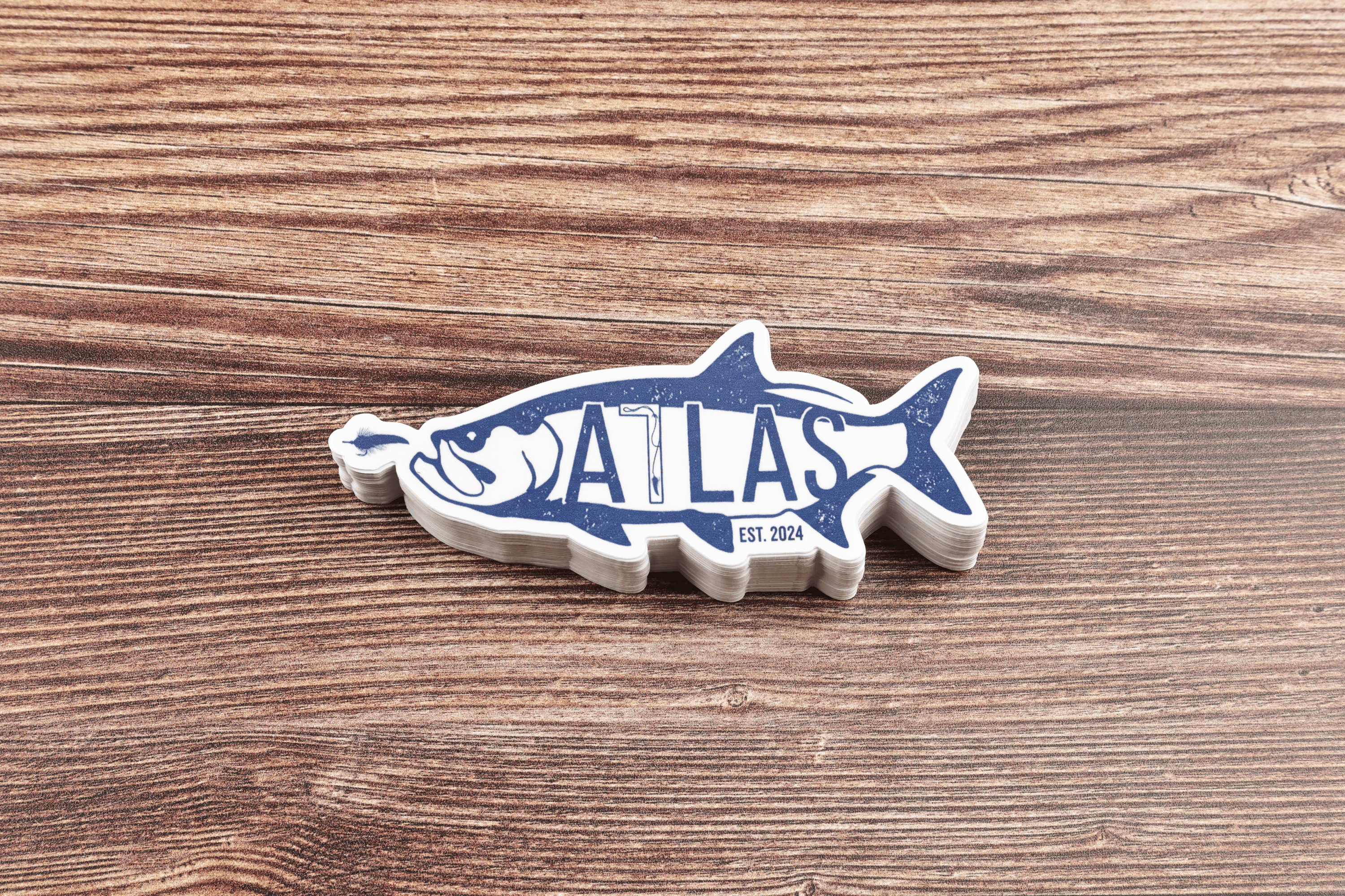 White vinyl tarpon-shaped sticker with distressed blue ATLAS logo and EST. 2024, stacked on brown wood-grain background