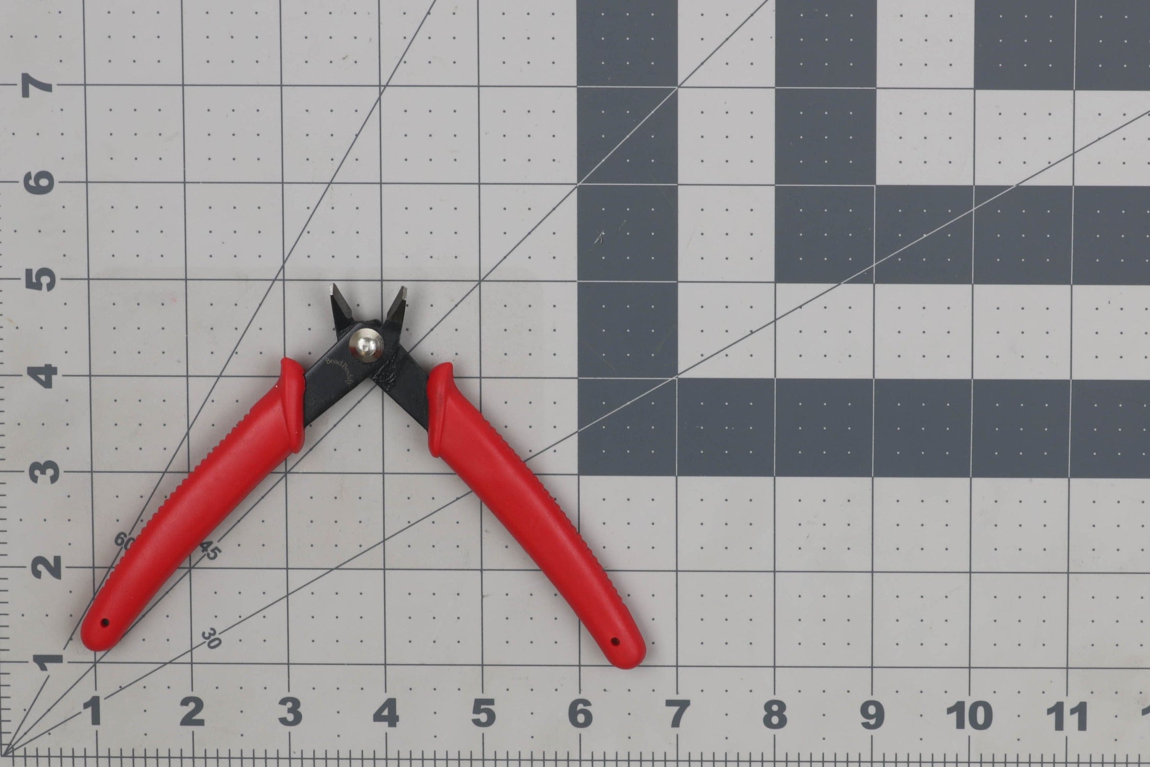 Red-handled Bead Buddy flush wire cutters with black metal jaws on gray gridded cutting mat, fly tying tool