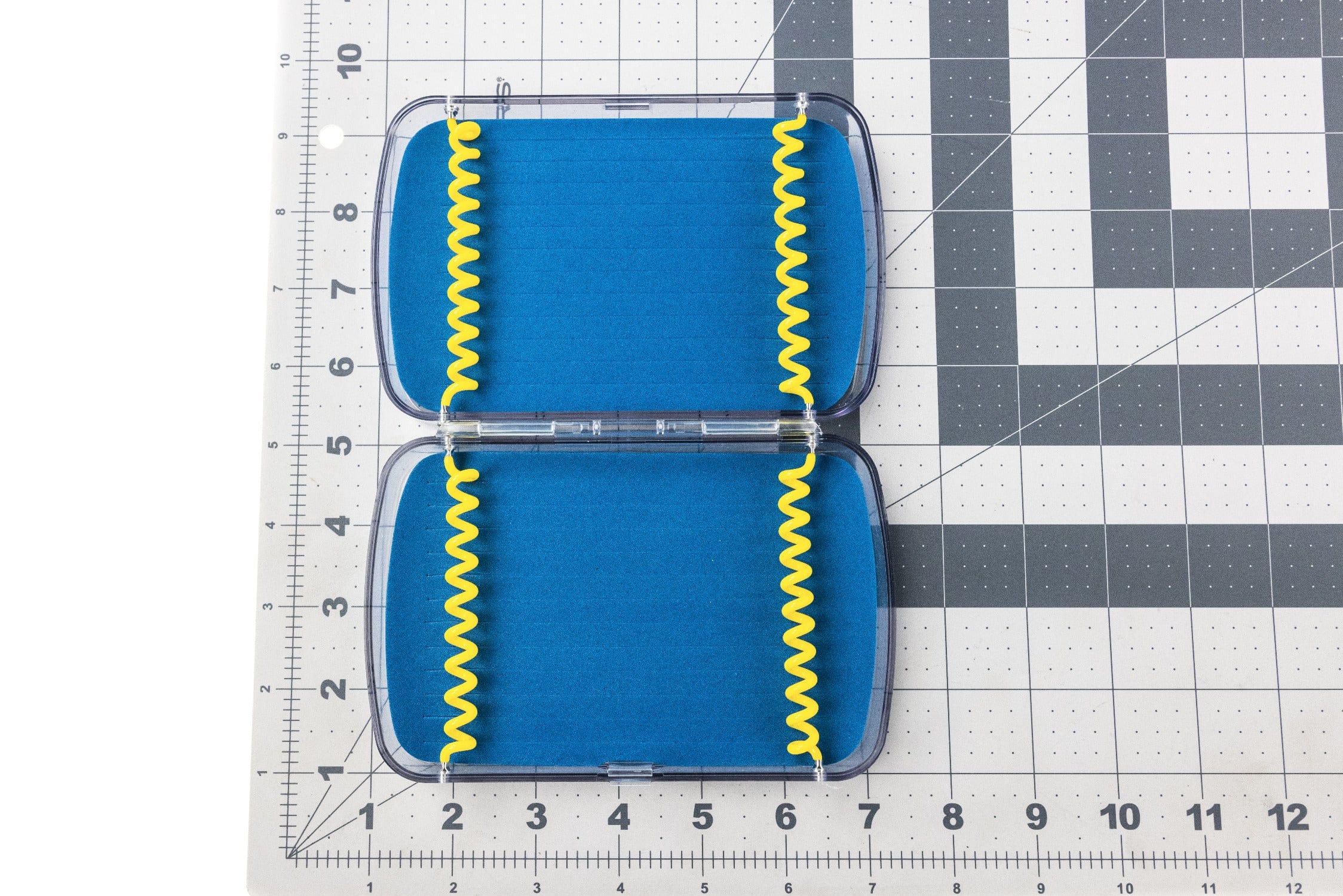 Clear hinged Cliff fly box with blue slit foam and yellow spiral retention cords on grid ruler backdrop