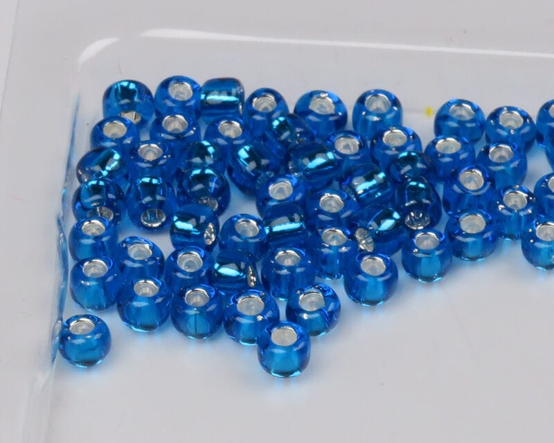 Transparent metallic-lined blue glass beads with silver centers for fly tying, scattered on white background