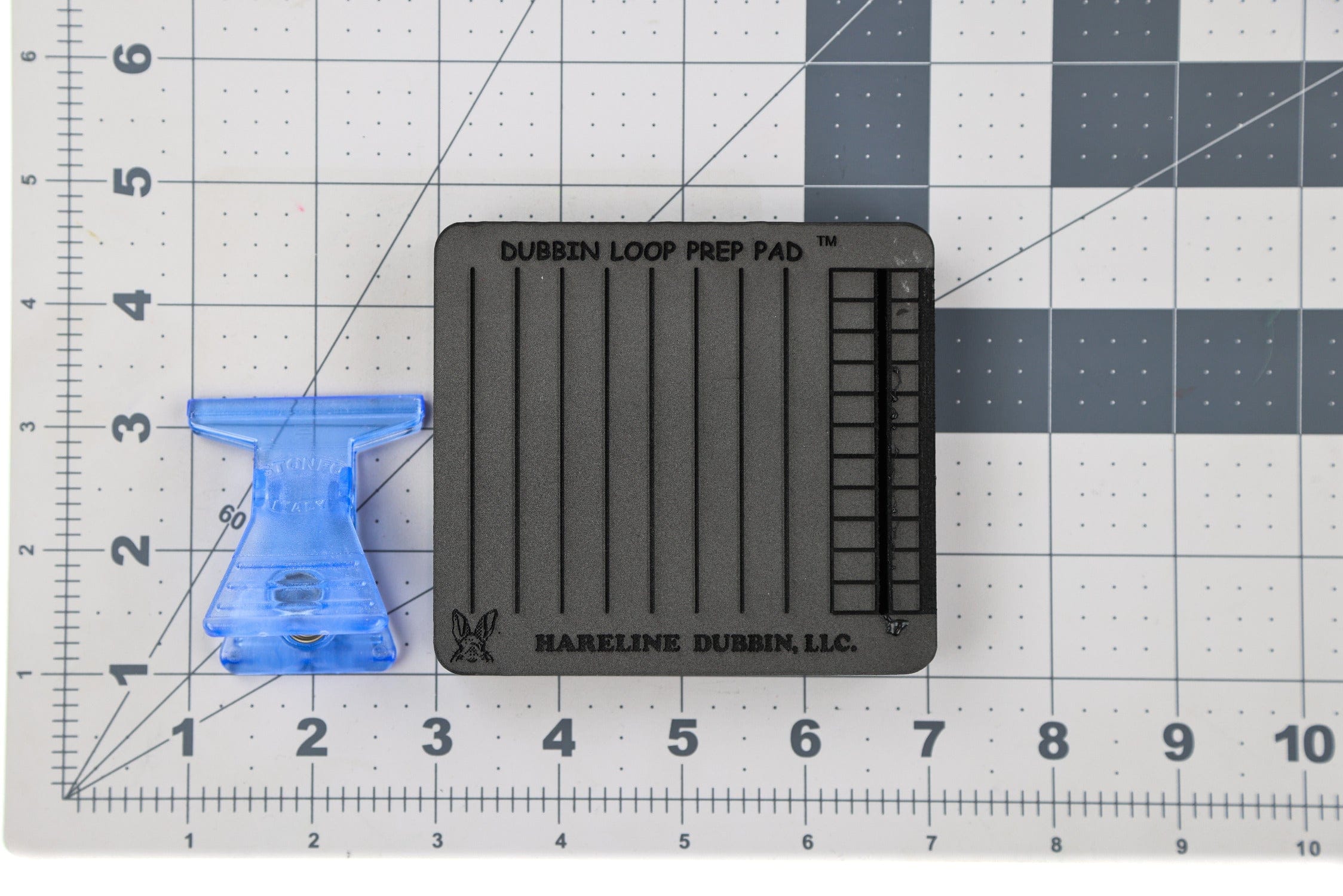 Black foam dubbing loop prep pad with vertical grooves and blue plastic clips on white gridded cutting mat, fly tying tool