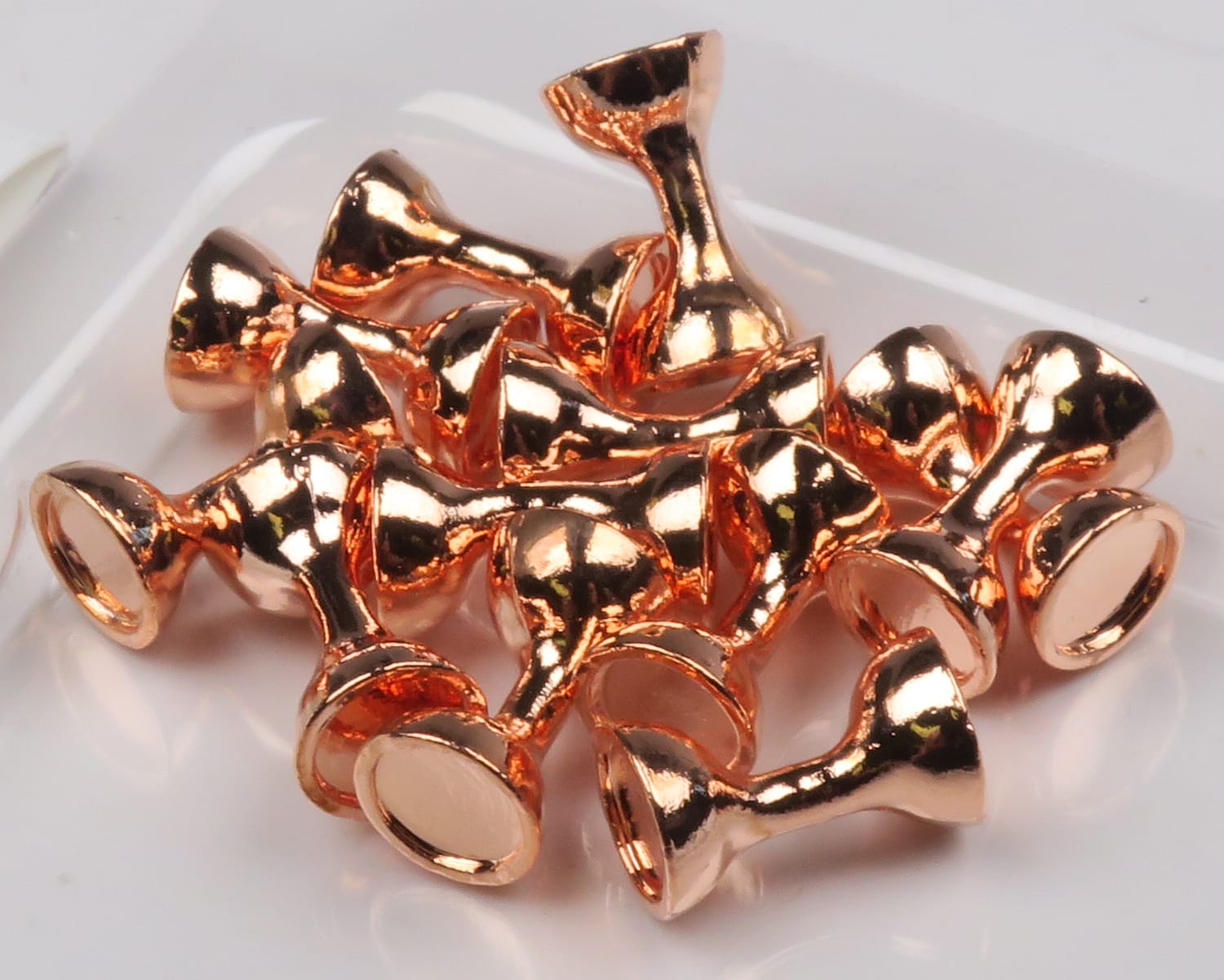 Copper-colored flat-end tungsten hourglass eyes for fly tying, shiny polished metal beads piled on white background