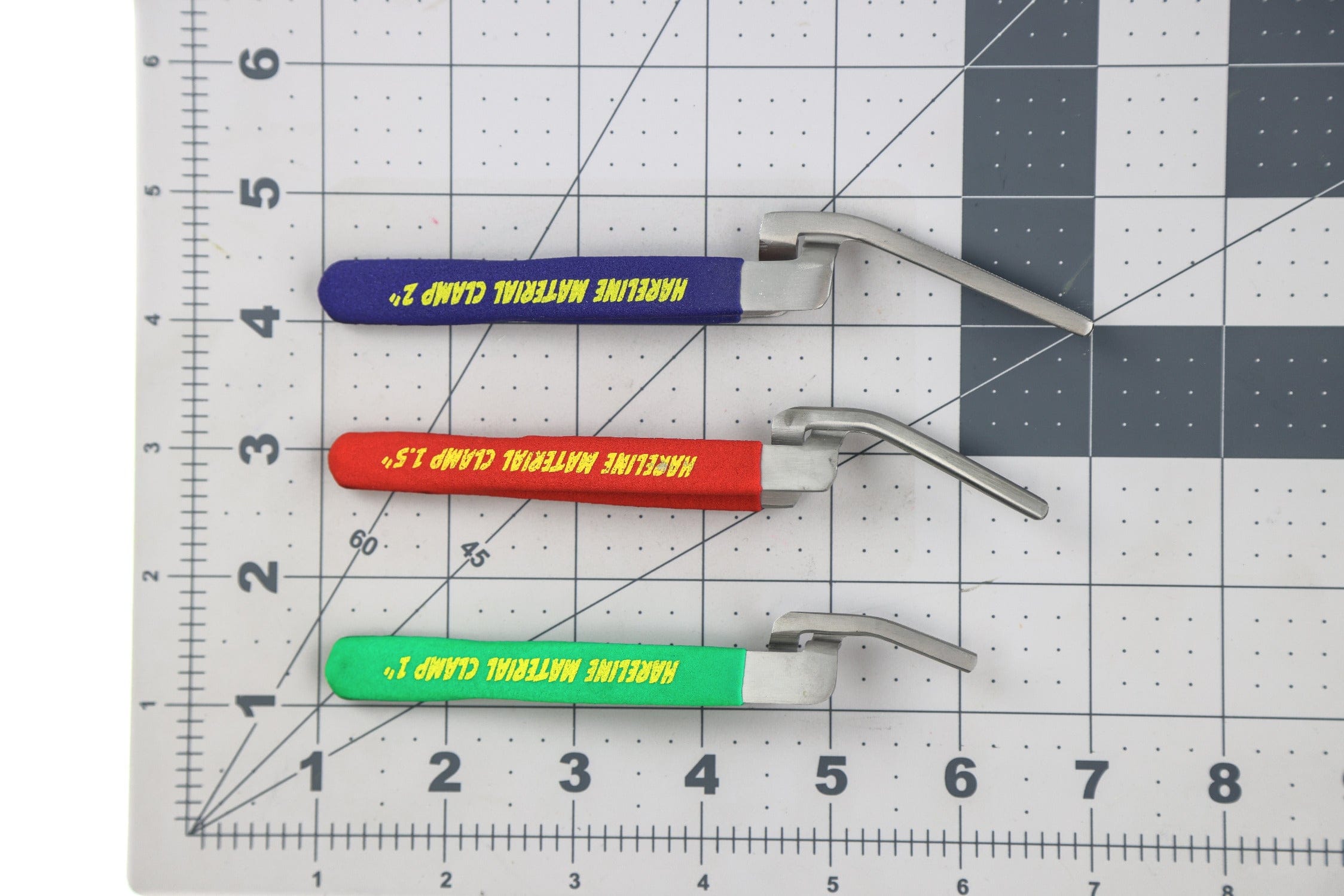 Three stainless steel material clamps with purple, red and green foam handles on gridded cutting mat, fly tying tool
