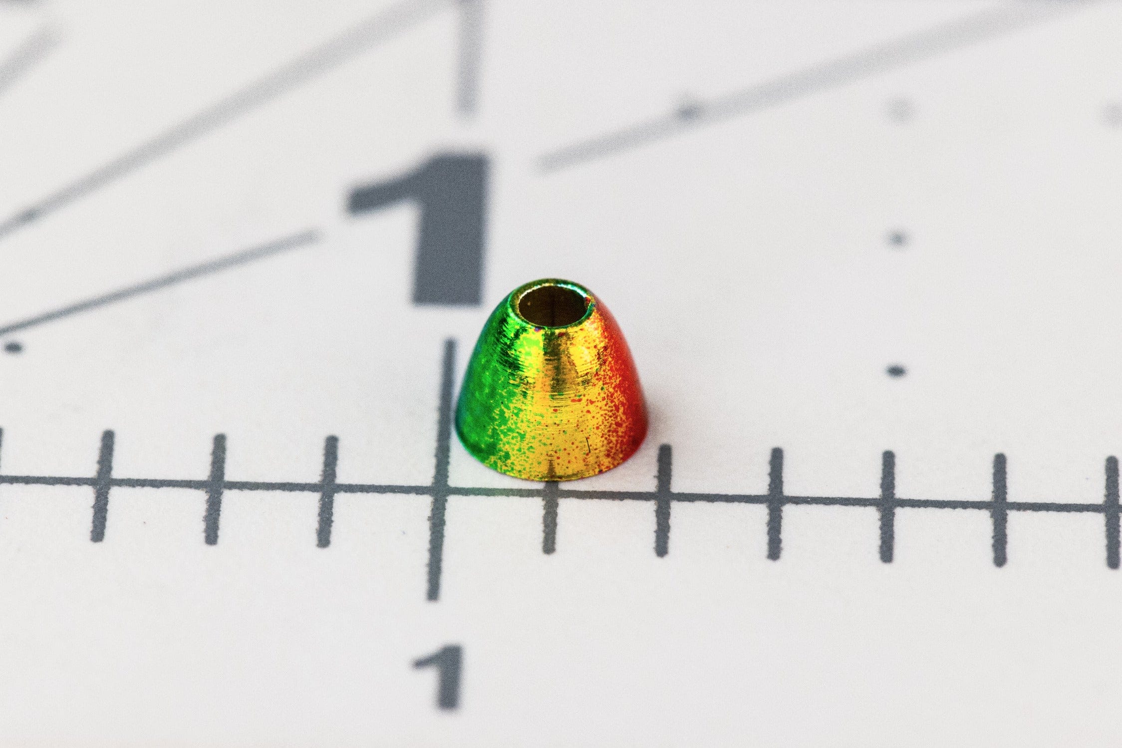 Rainbow iridescent brass cone in green-yellow-red finish, small fly tying cone placed on ruler grid