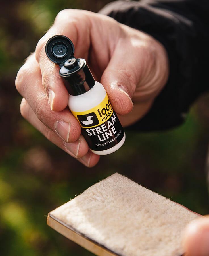 Hand holding white Loon Stream Line treatment bottle with black flip cap and yellow label over beige wool pad, fly fishing