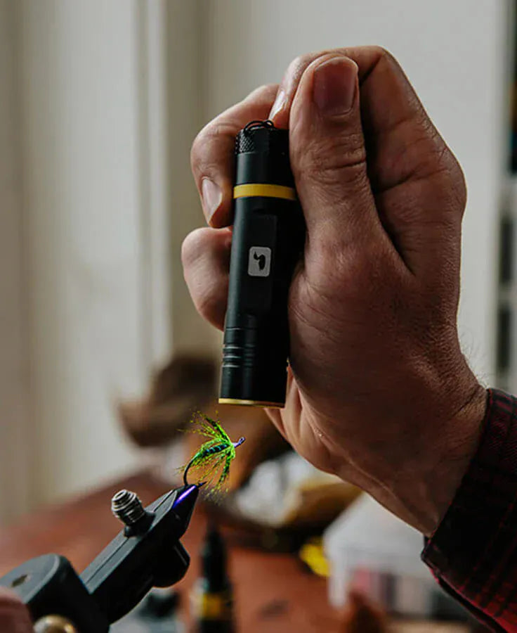 Hand holding black Loon UV bench light with yellow trim illuminating green synthetic fly in vise for fly tying