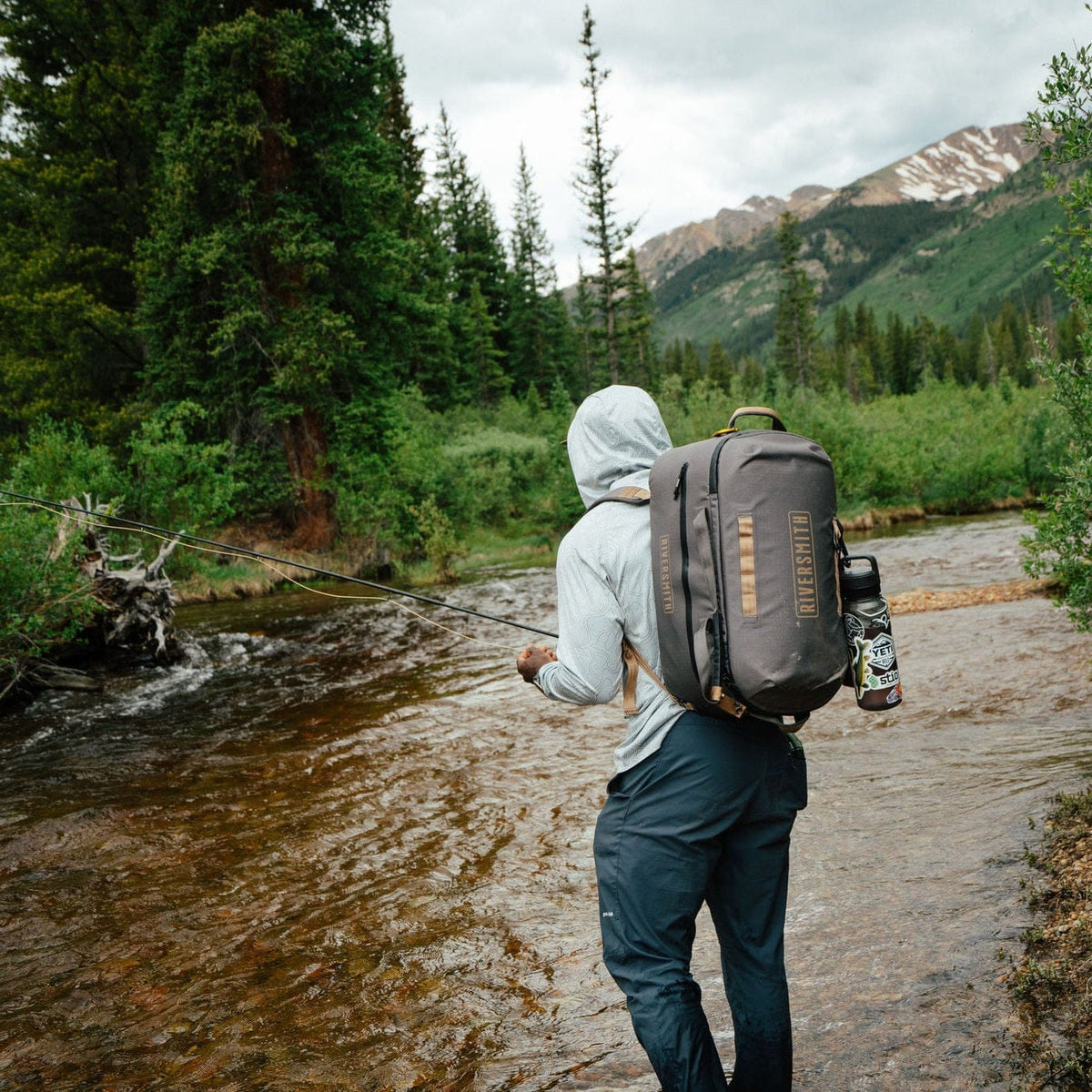 Angler wearing gray Riversmith waterproof duffel backpack while fly fishing in mountain river valley