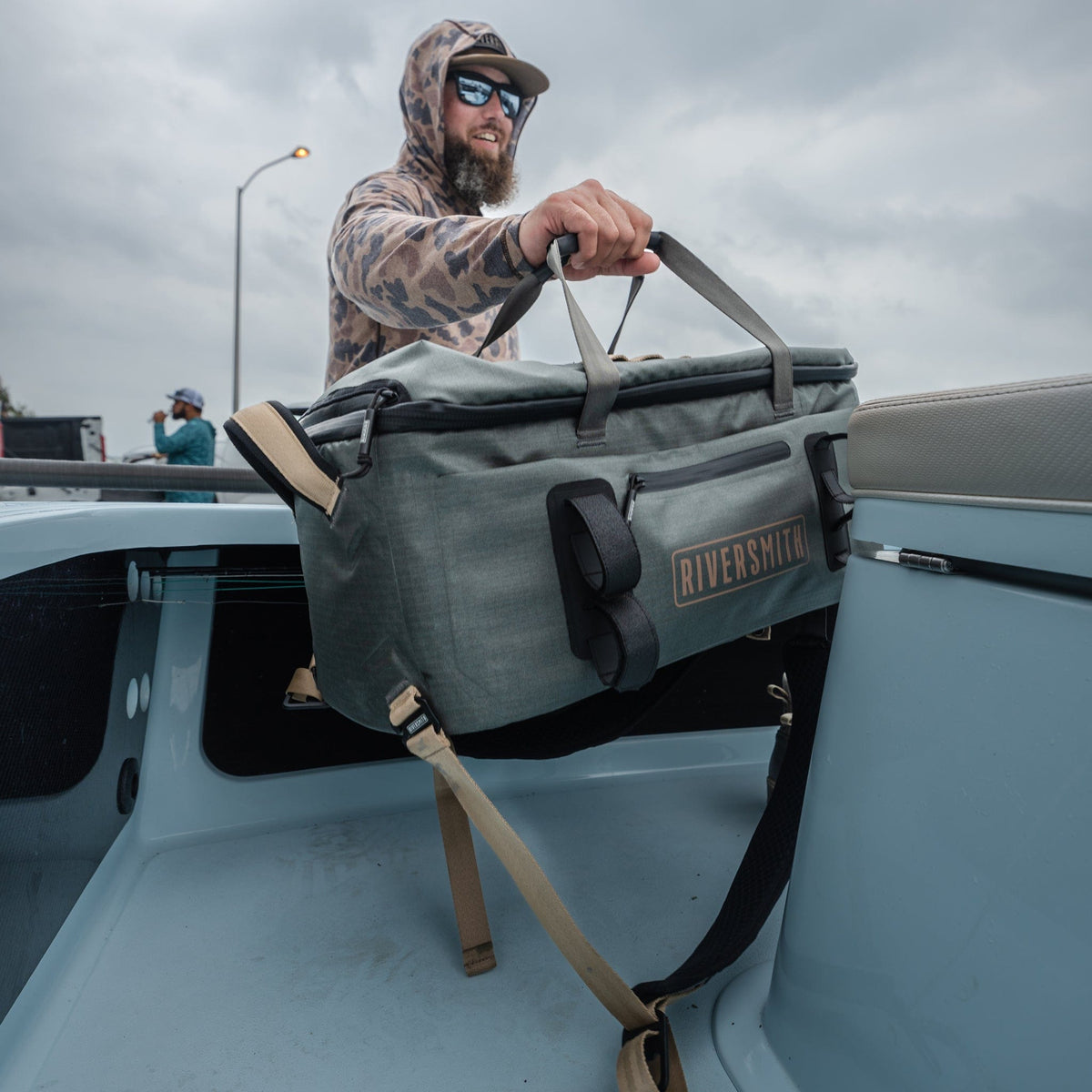 Riversmith waterproof duffel bag in gray being held over truck bed with tan handles and shoulder strap