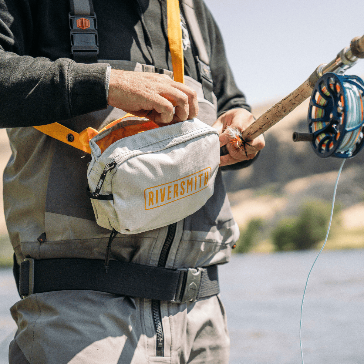Riversmith PackOut fly fishing waist pack in cream with orange accents, worn by angler holding fly rod near water