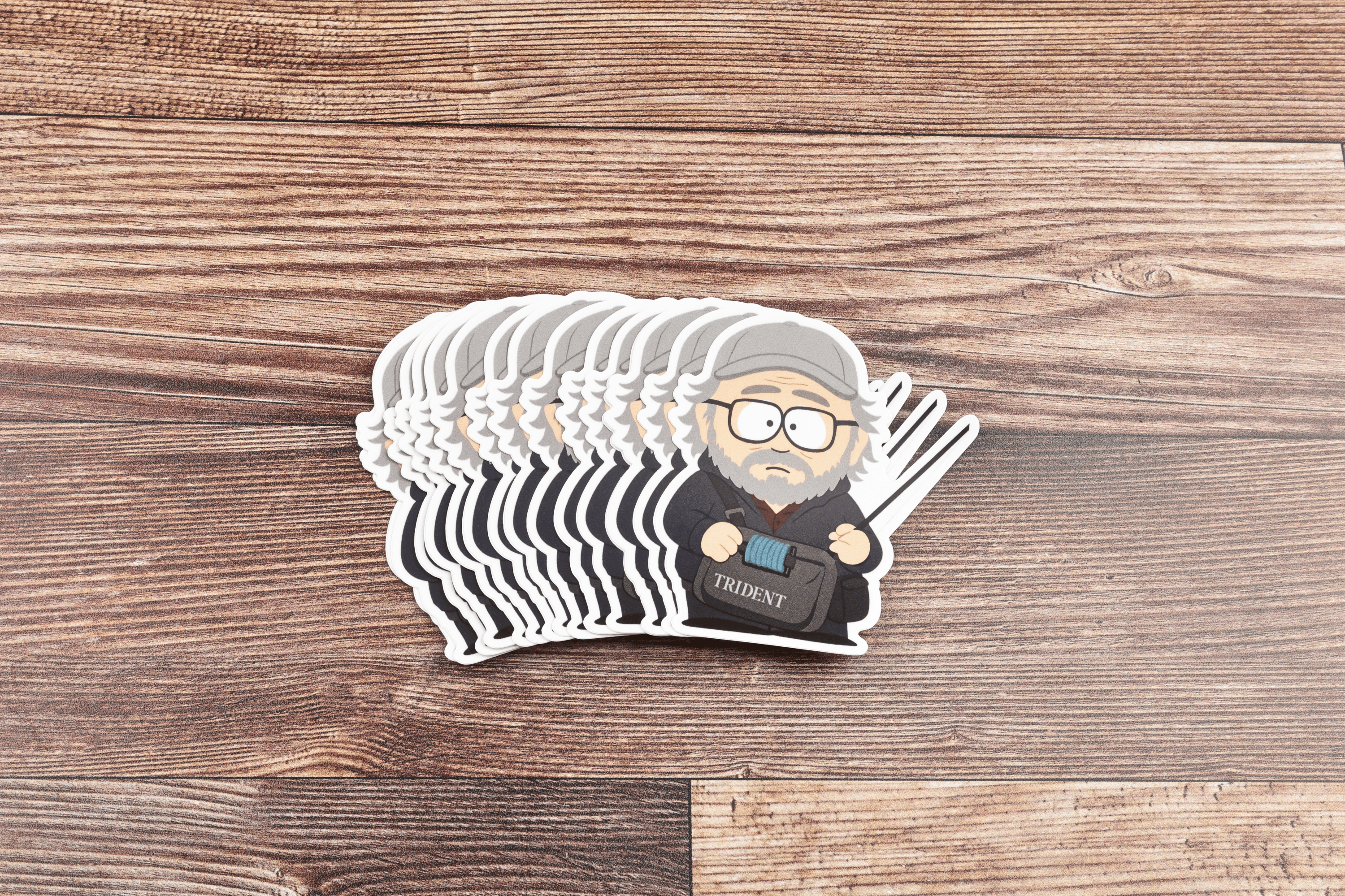 Stack of die-cut Rom Rosenbauer stickers showing cartoon bearded man with gray cap, glasses and Trident bag on wood