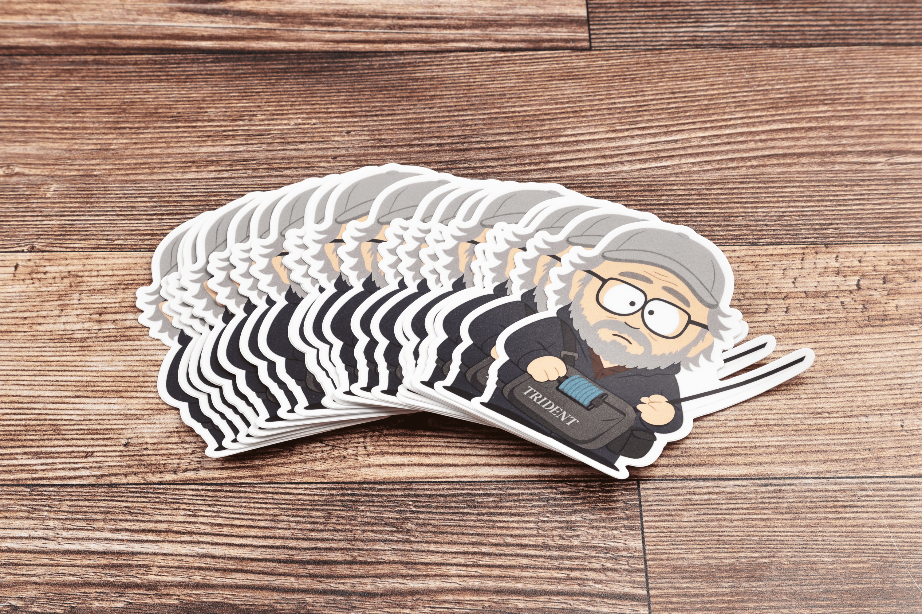 Die-cut vinyl sticker stack showing cartoon Rom Rosenbauer, gray-bearded man in gray beanie holding a Trident bag on wood