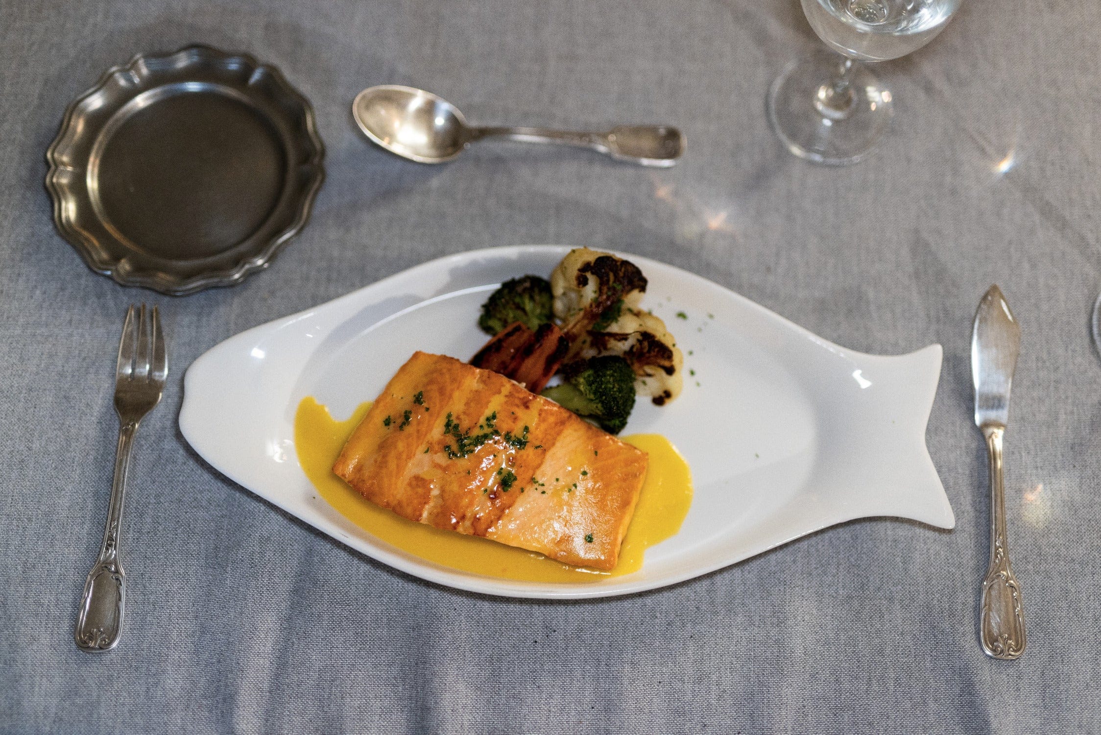 Seared salmon on white fish-shaped ceramic plate with yellow sauce and roasted broccoli on gray linen, Trident