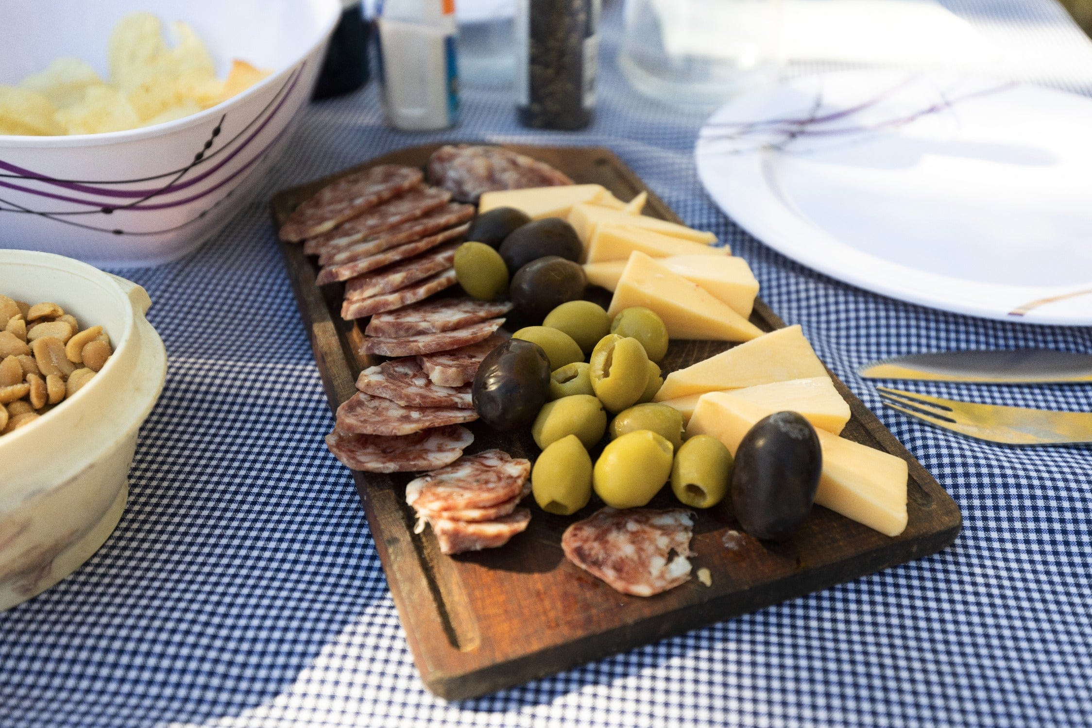 Wooden charcuterie board with sliced marbled salami, green and black olives, yellow cheese wedges on blue gingham picnic