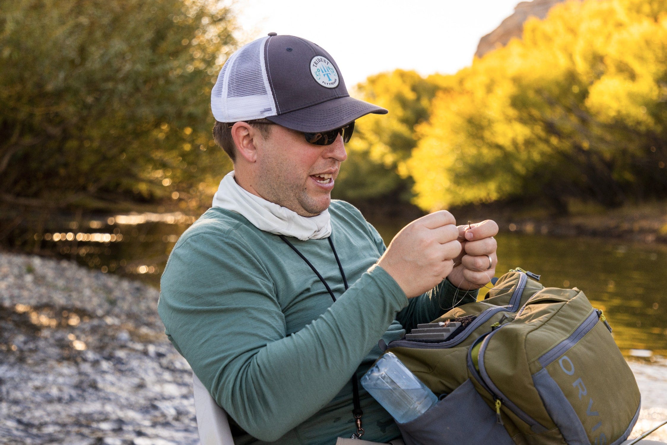 Fisherman wearing gray mesh Trident hat and green shirt, tying fly line by river with olive backpack on Chimehuin Safaris
