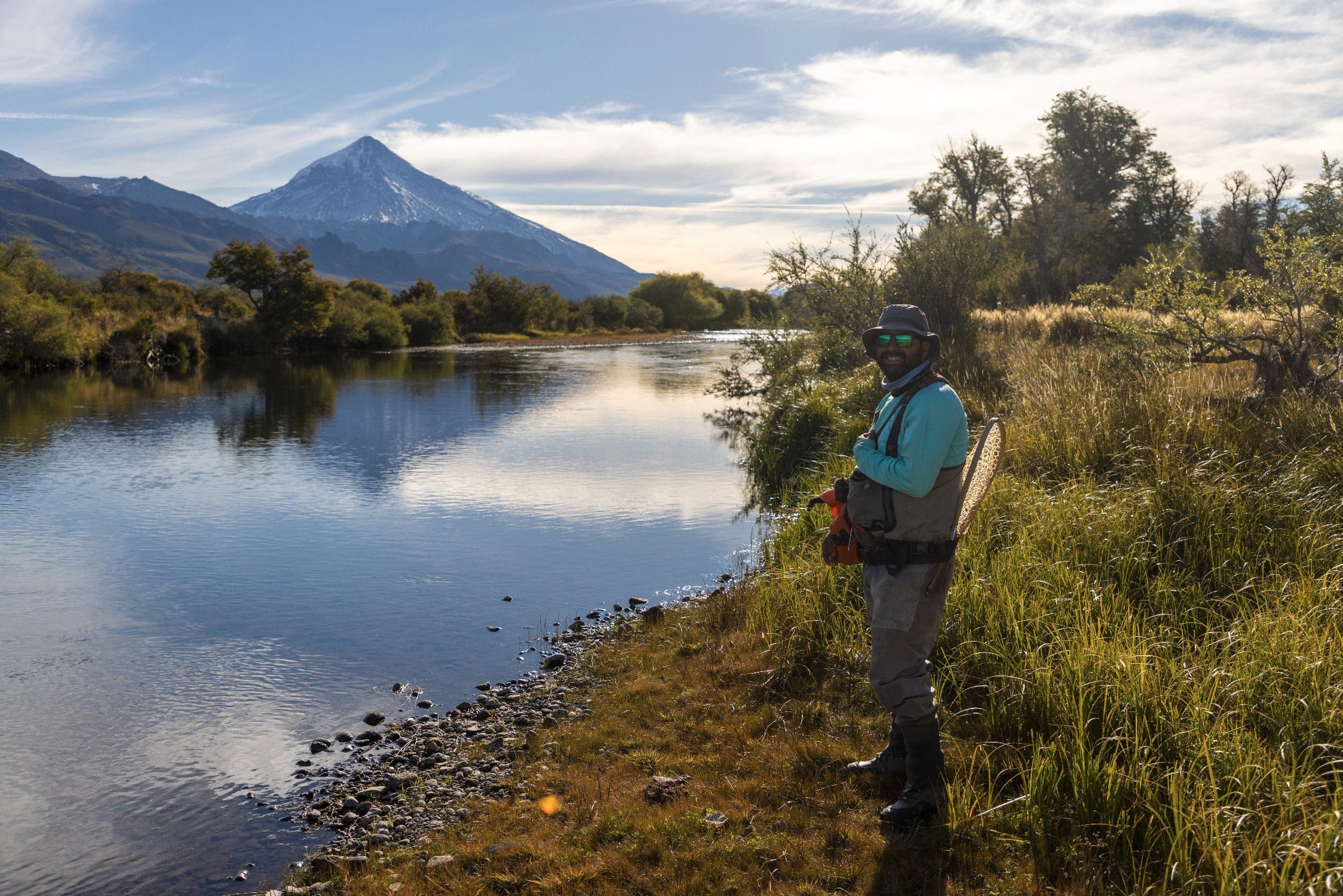 Angler in teal shirt, bucket hat and waders on golden grass riverbank with wooden net, blue river and snow-capped peak,