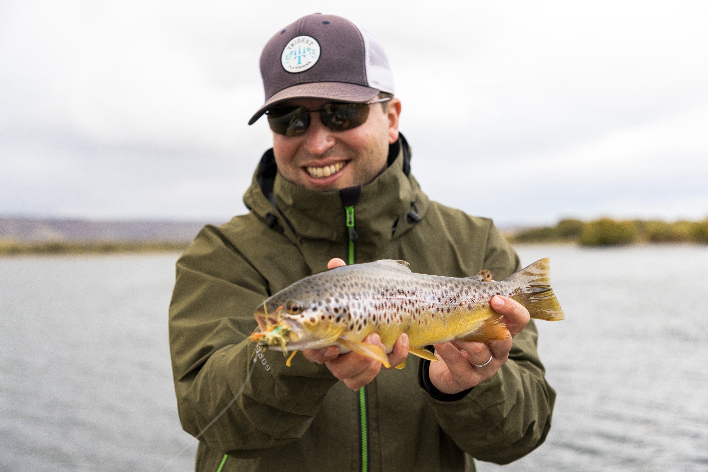 Smiling angler in olive green Gore-Tex jacket and brown Trident cap holding a speckled brown trout on a Chimehuin safari