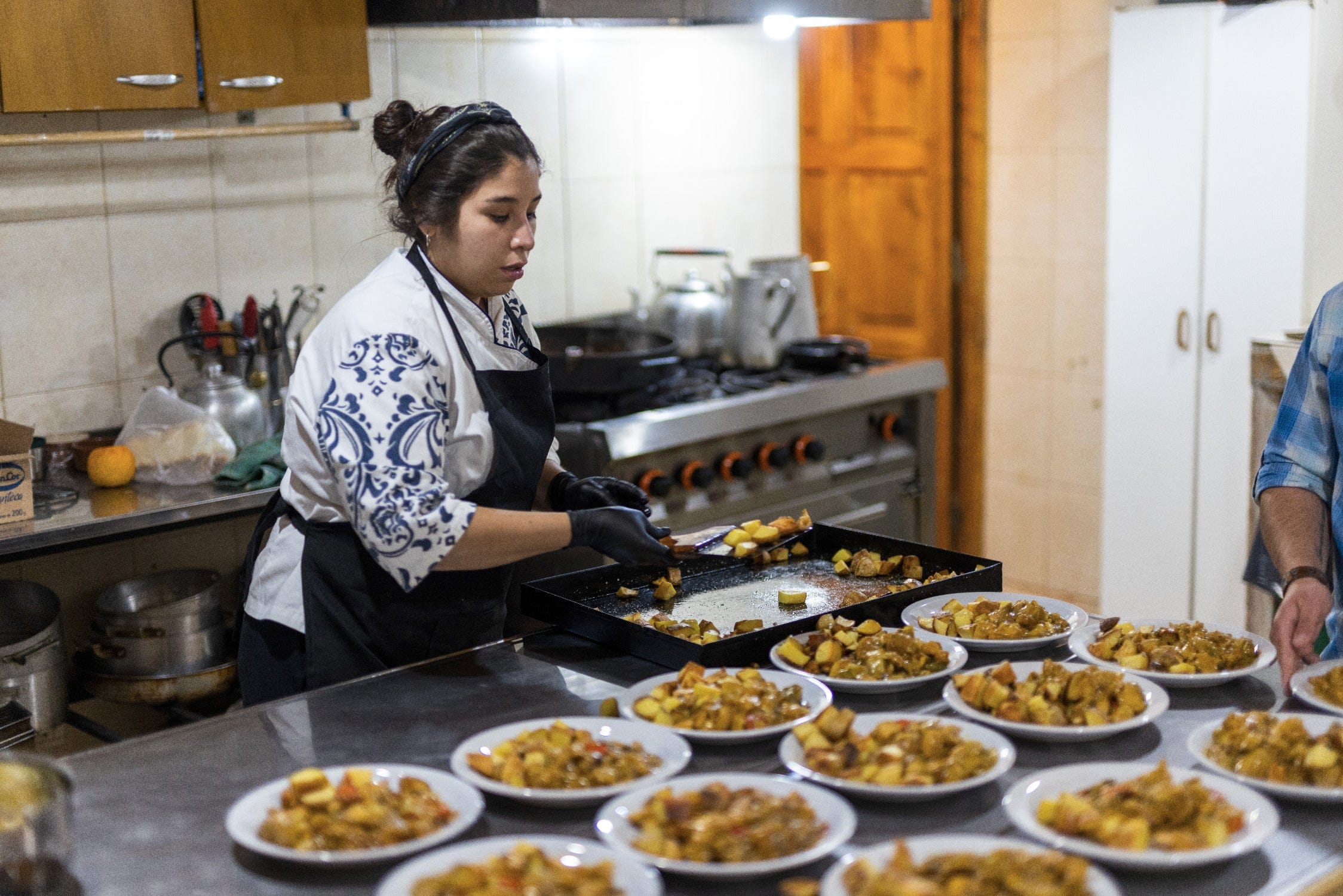 Woman chef in black apron plating roasted potatoes on white plates in a stainless-steel lodge kitchen