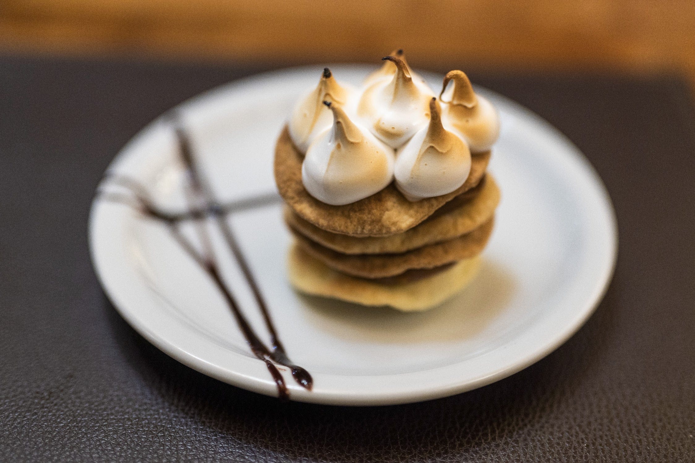 Stacked golden-brown cookie dessert topped with toasted white meringue peaks on a white plate with chocolate drizzle