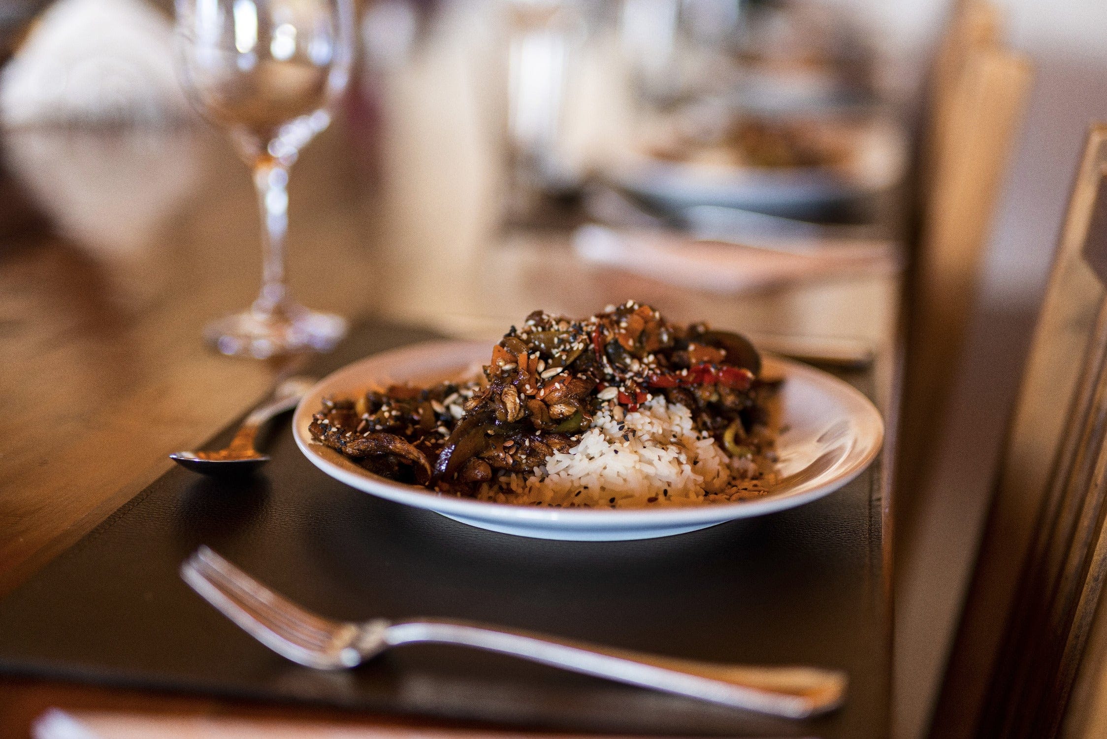 White plate of rice and sesame-topped brown stir-fry on a wooden lodge dining table with blurred wine glass