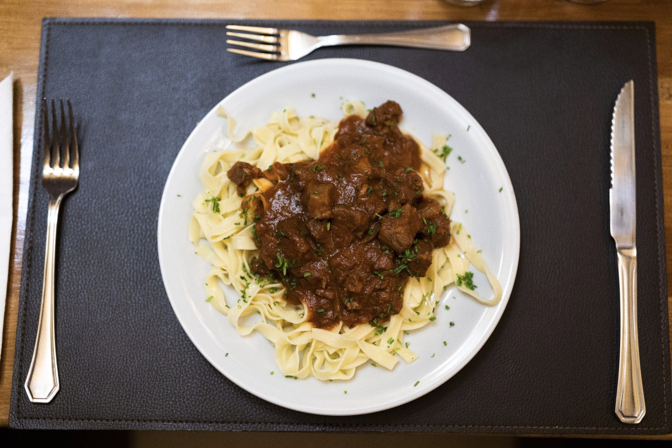 Plate of tagliatelle with brown beef ragout and chopped parsley on black leather placemat with metal cutlery, lodge dining