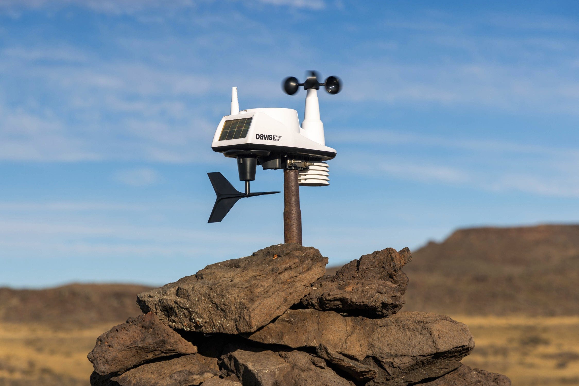 White weather sensor with spinning black cups and wind vane on rusted pole on rocks under blue sky Jurassic Lake Lodge