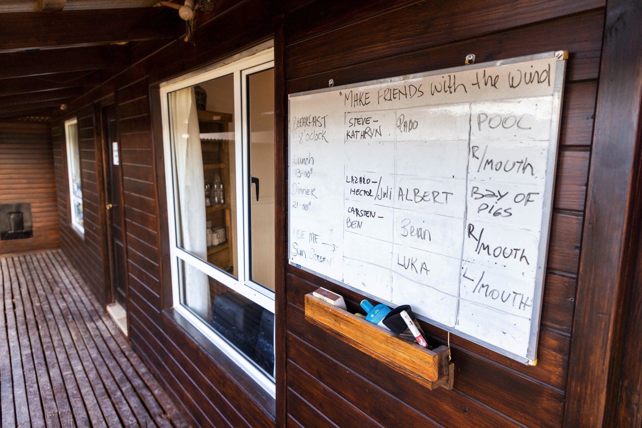 Dark brown wooden lodge porch with sliding glass window and written whiteboard schedule above a rustic timber deck, trip