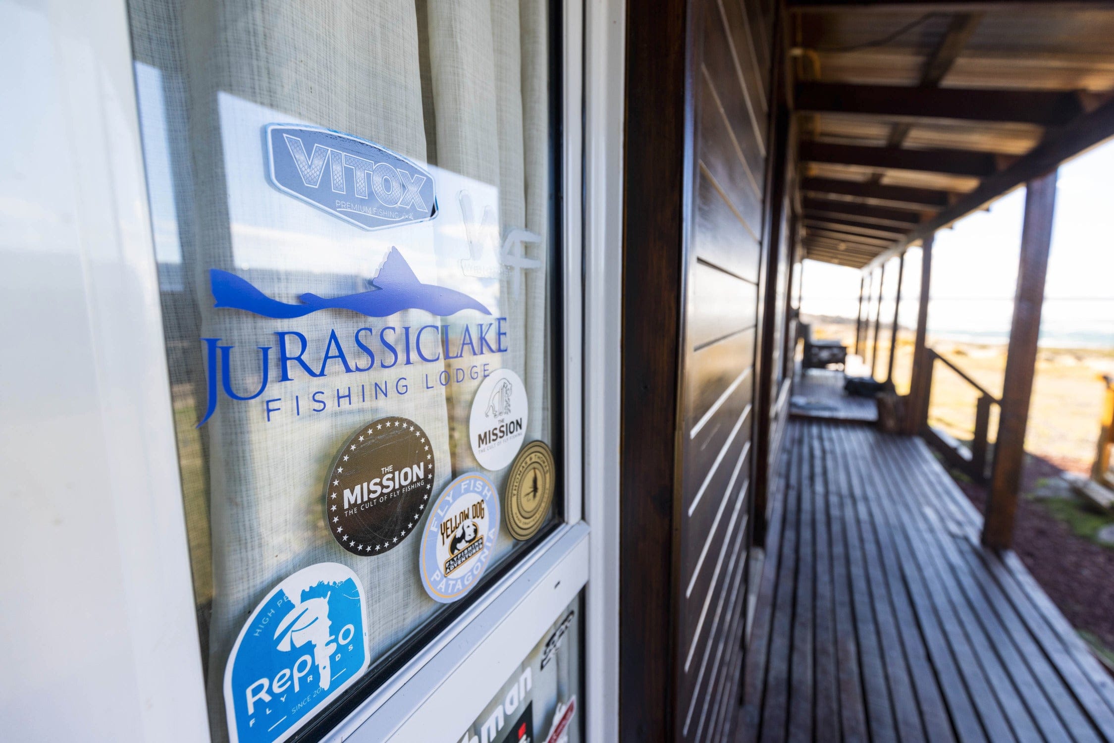 Glass door with blue Jurassic Lake lodge decal and stickers, sheer curtain and rustic wooden veranda overlooking shore