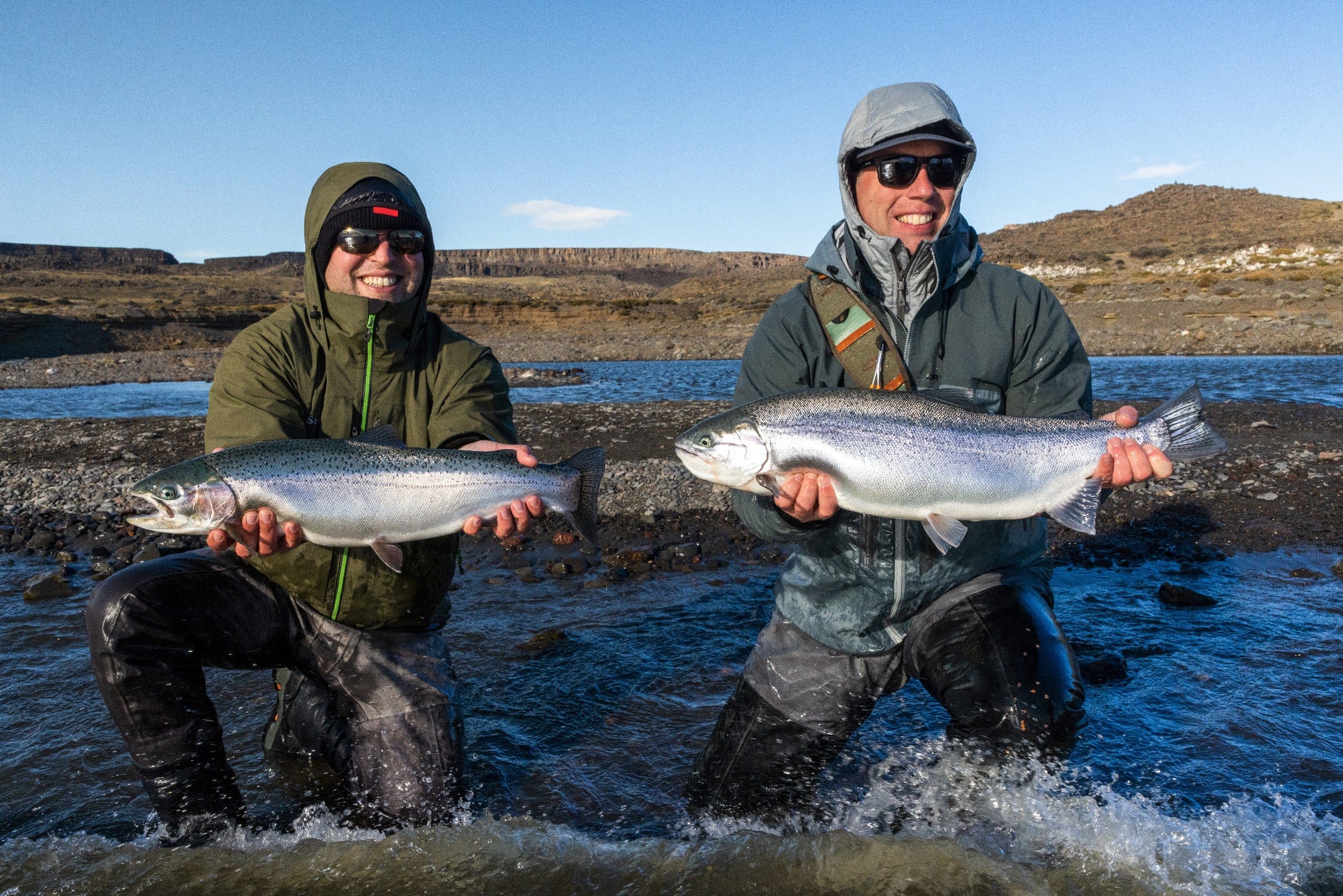 Two anglers in green and gray waterproof jackets holding large silver trout in blue river at Jurassic Lake Lodge fishing