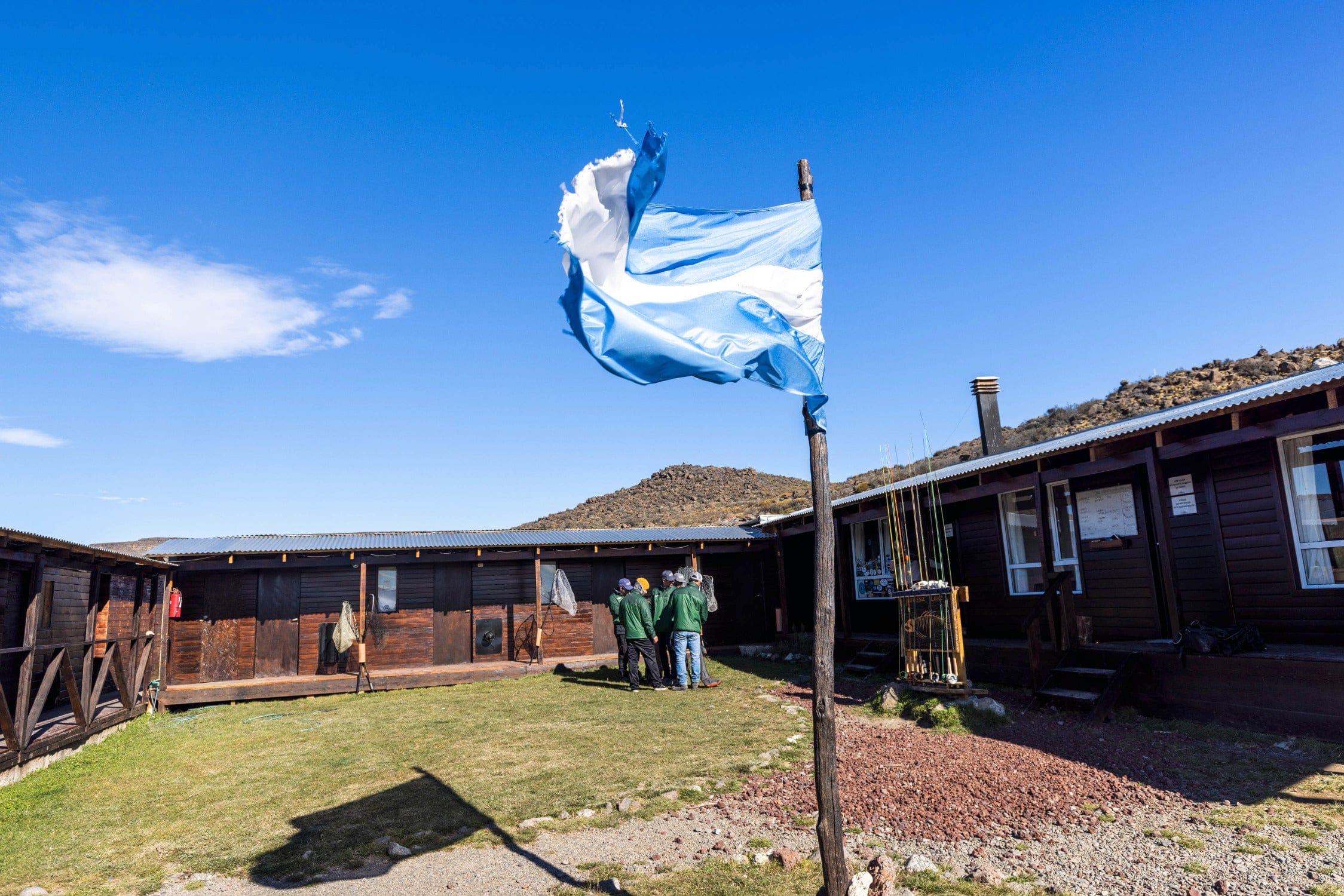 Torn blue-and-white flag on a wooden pole above dark timber cabins with corrugated metal roofs at Jurassic Lake Lodge trip