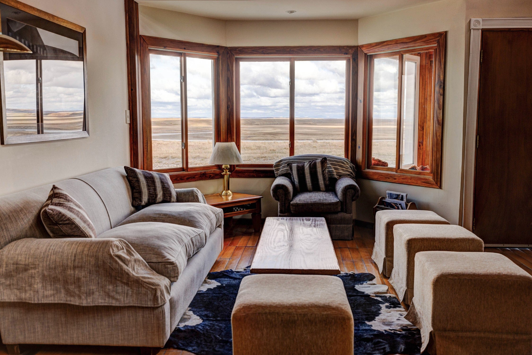 Cozy beige fabric sofa and armchair in lodge living room with warm wood window frames, brass lamp and panoramic prairie view