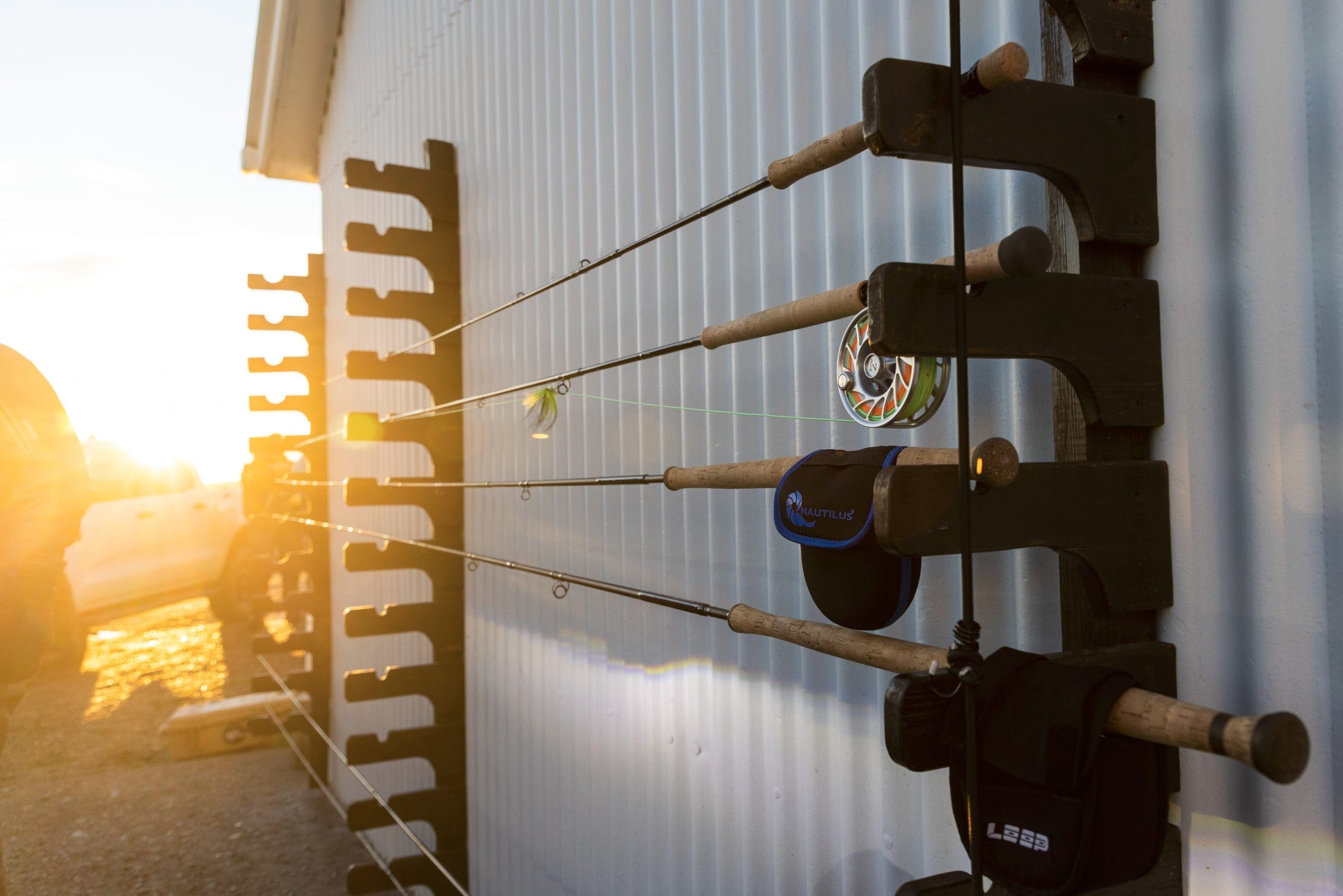 Wooden rod rack holding cork-handle fly fishing rods with metal reels against white corrugated wall at sunset