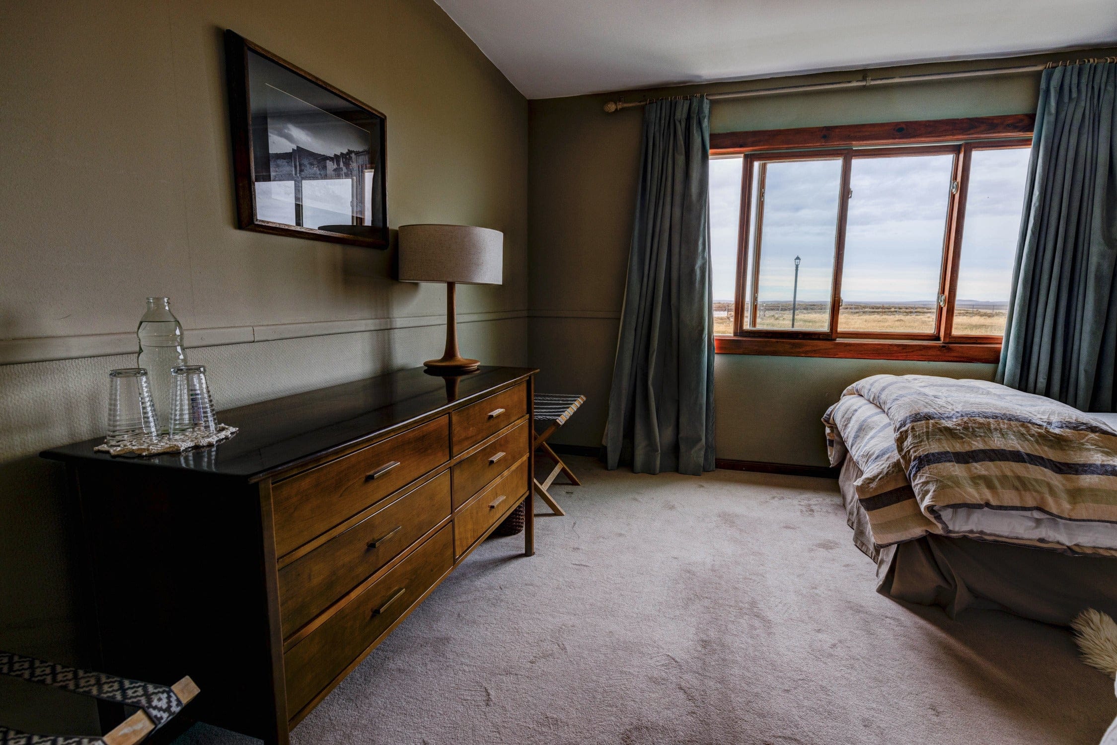 Cozy lodge room with wooden dresser, beige linen lamp, teal curtains and striped bed linens by a window with plains view