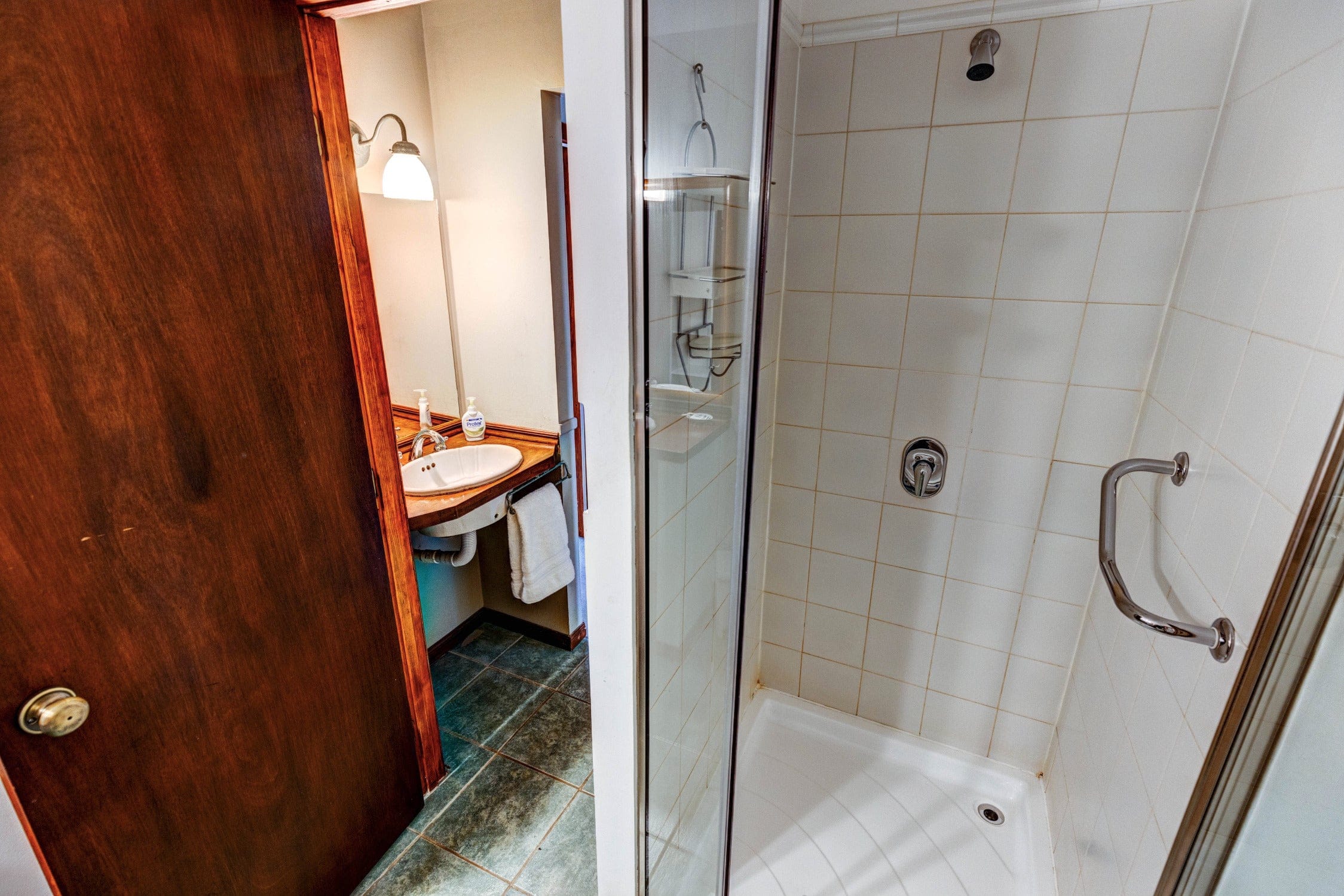 Compact lodge bathroom with white tiled shower, glass door and chrome grab bar, wood vanity with white sink and green slate