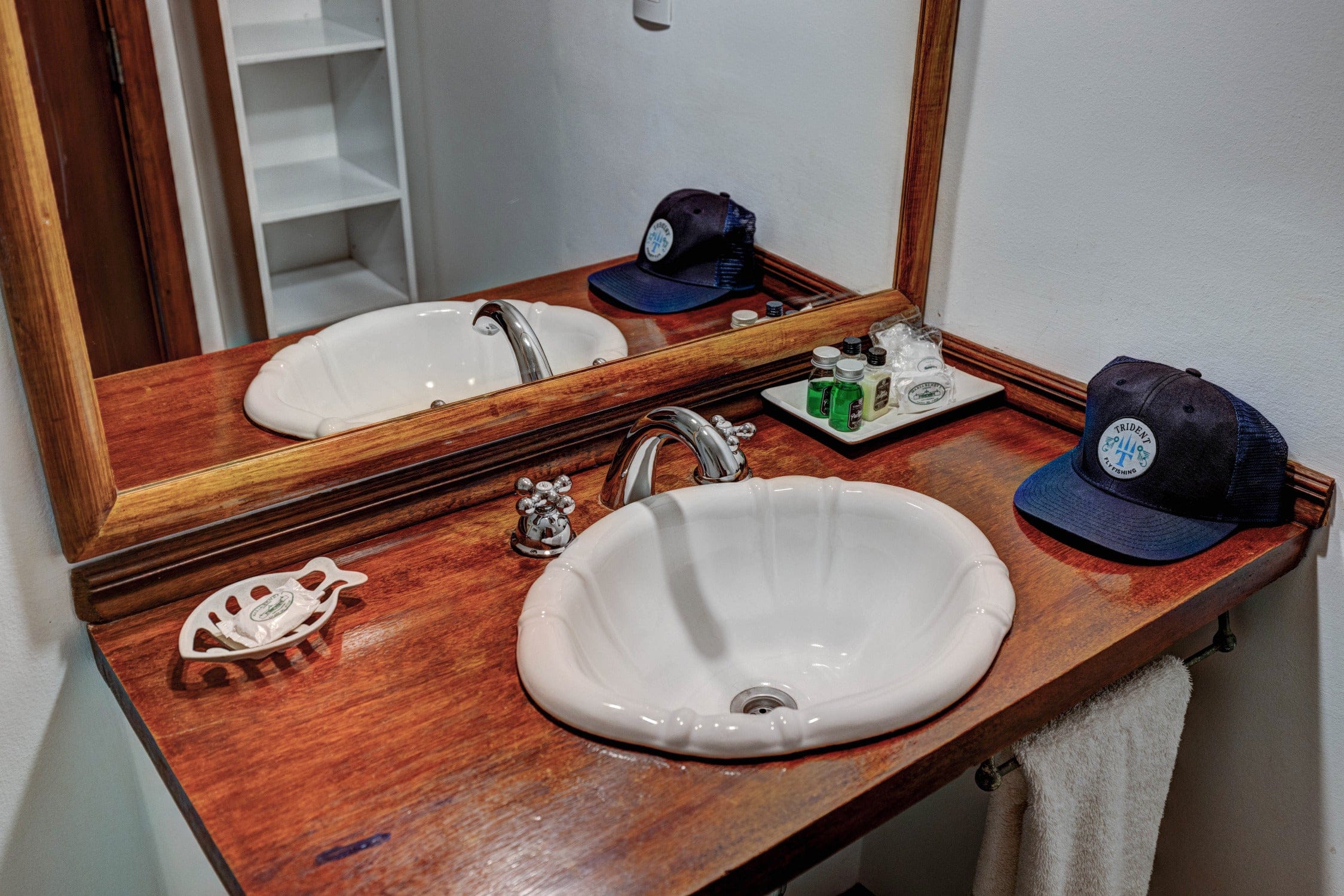 Wooden vanity with white scalloped ceramic sink, chrome faucet, navy Trident cap and toiletry bottles in lodge bathroom