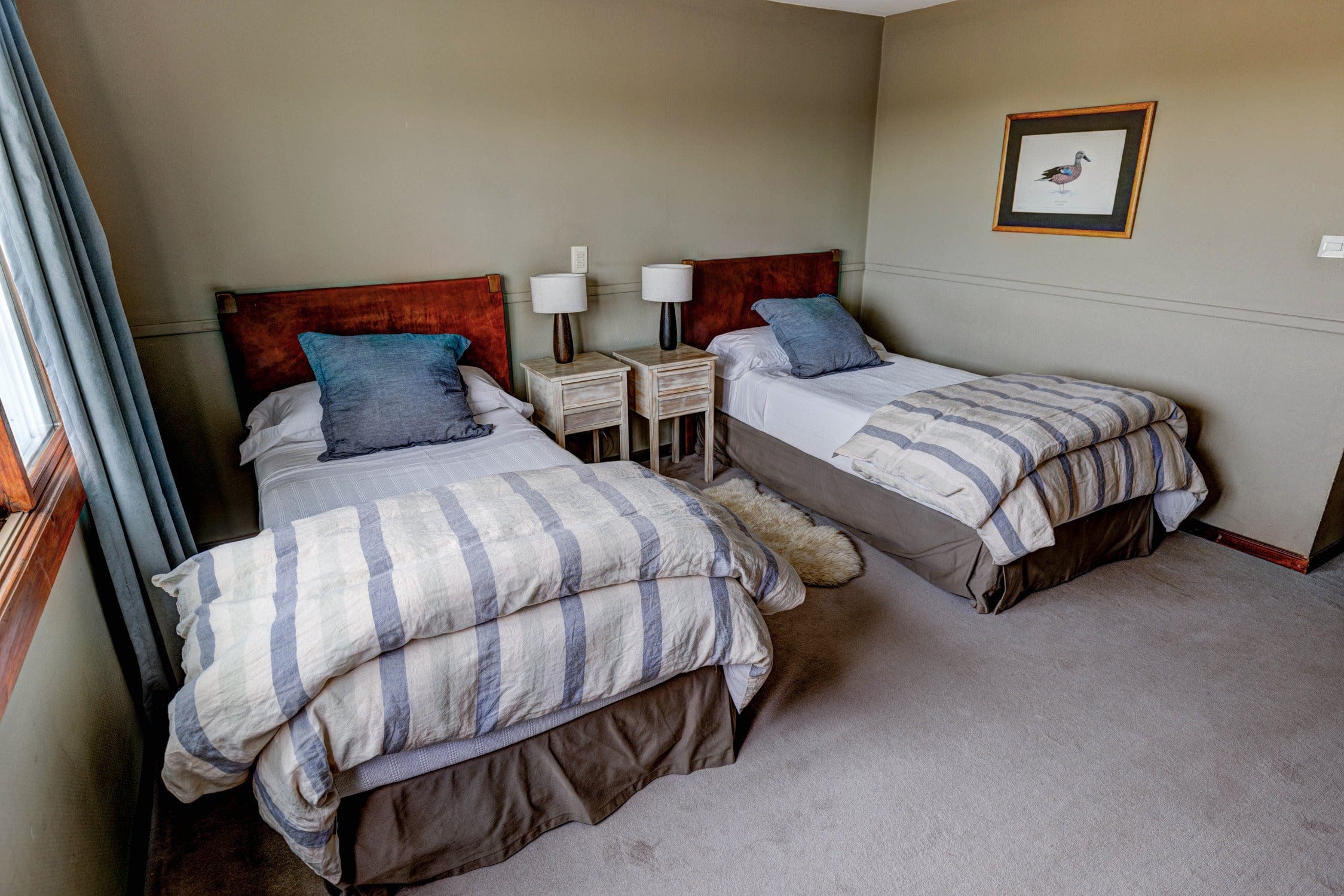 Cozy lodge bedroom with two twin beds, striped blue-gray duvets, wooden headboards, bedside tables and fabric lamps