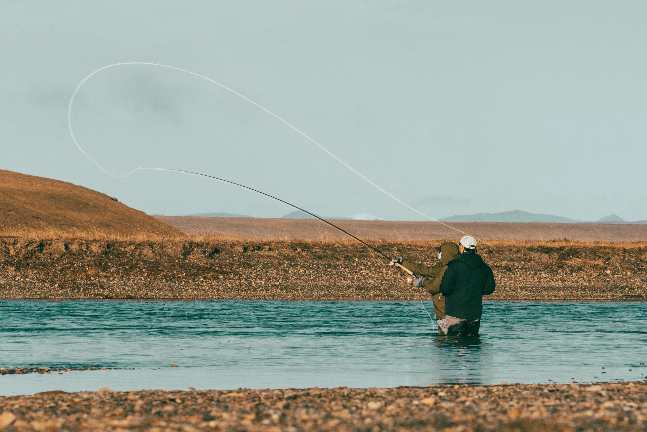Two anglers in waders fly-fishing in teal river with looping white cast and brown grassy shoreline, fishing trip