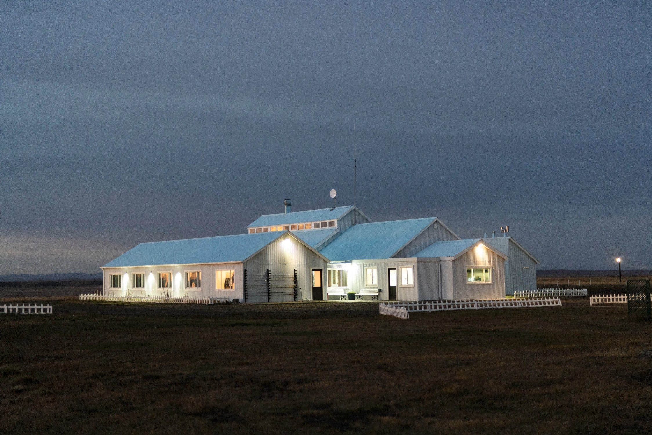 White corrugated metal lodge with blue gabled roofs, warm lit windows and picket fences at dusk, accommodation