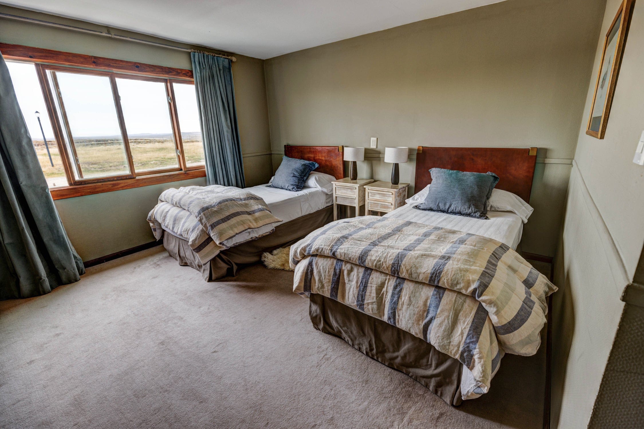 Twin beds with striped beige-blue quilts, wooden headboards and bedside lamps in a cozy rustic lodge room with window view