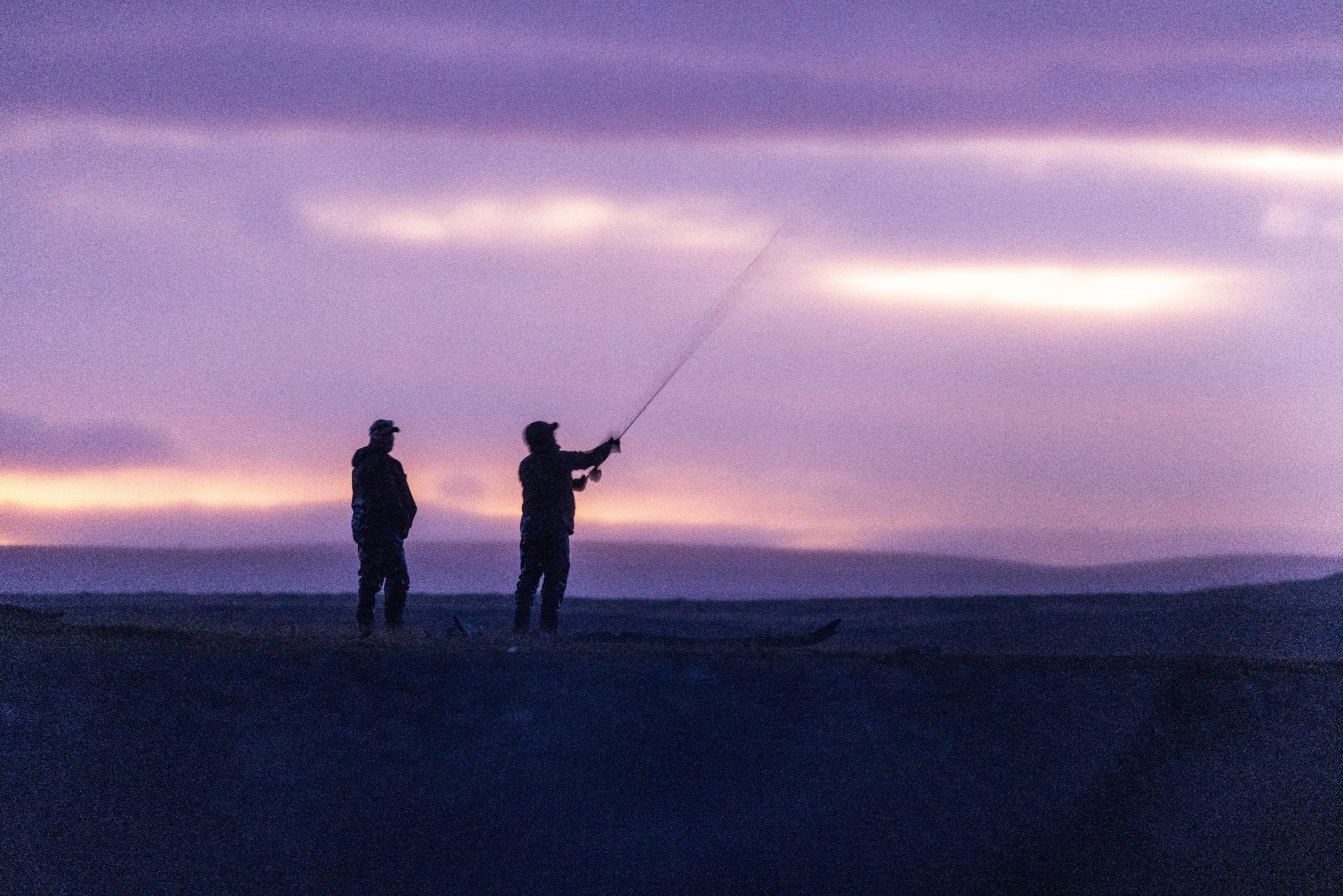 Two silhouetted fishermen casting on a dark shore under a purple-pink sunset, fishing trip near lodge