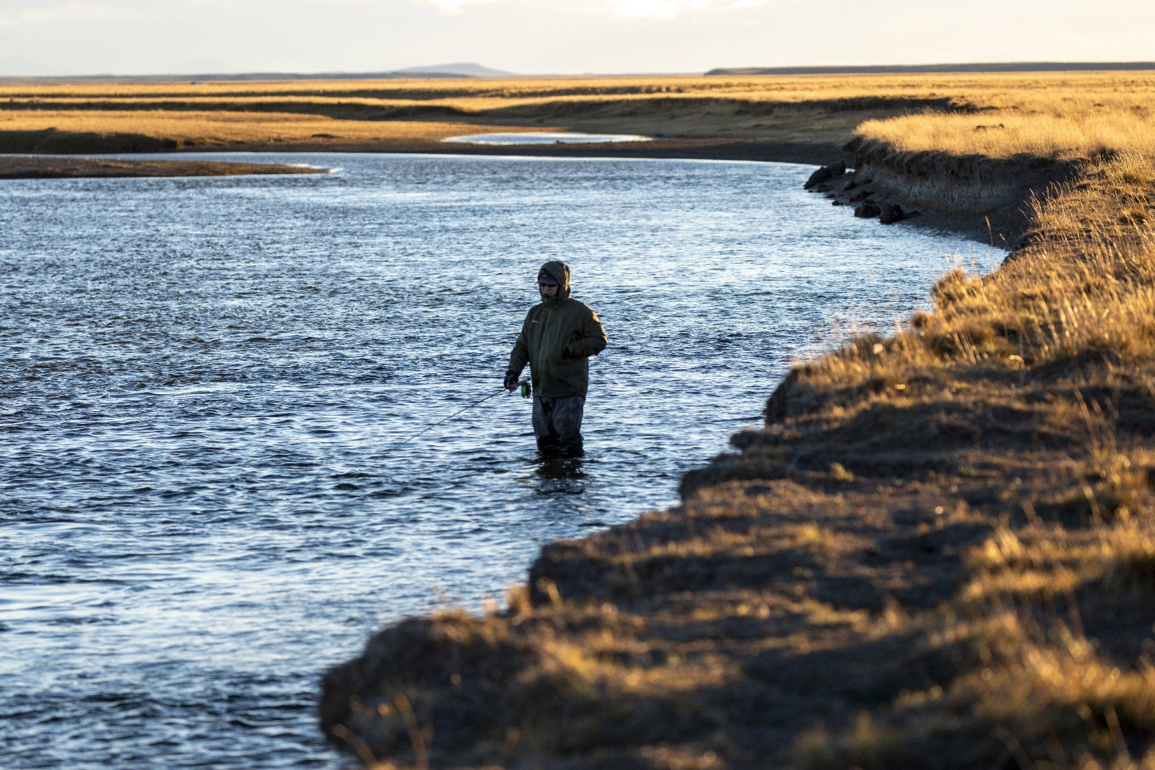 Angler in green hooded jacket wading in blue river beside golden grassy banks at sunset on a fishing trip