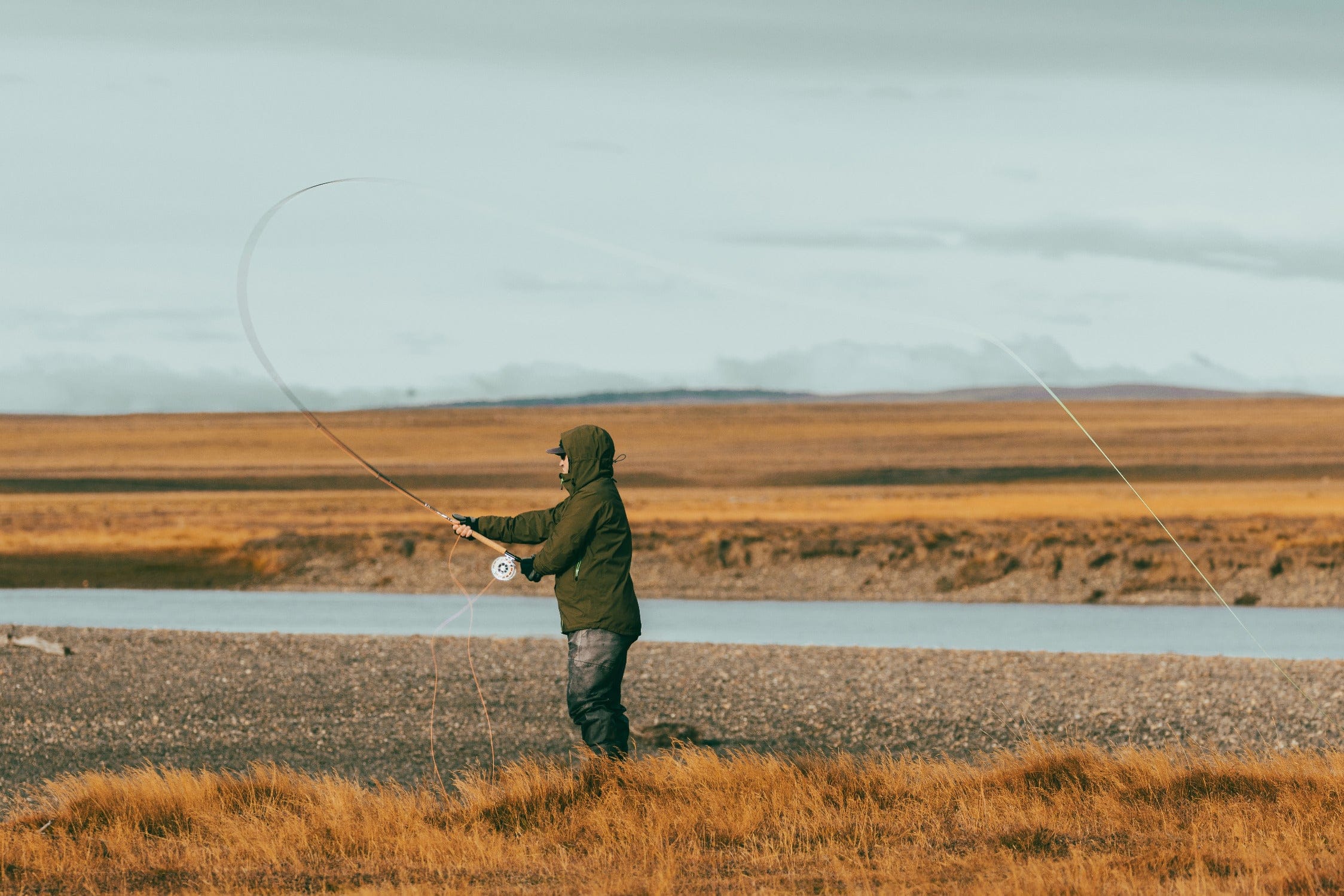 Angler in olive-green hooded jacket fly fishing on a rocky riverbank with golden grasses on a wilderness trip