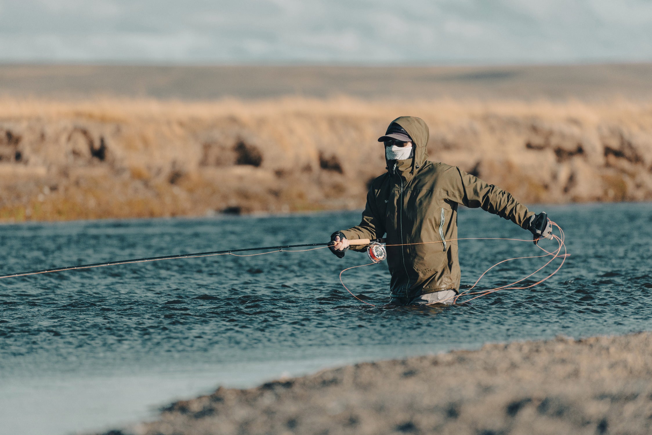 Angler in olive green hooded jacket and waders fly-fishing waist-deep in blue water, mask and sunglasses, Maria Behety Lodge