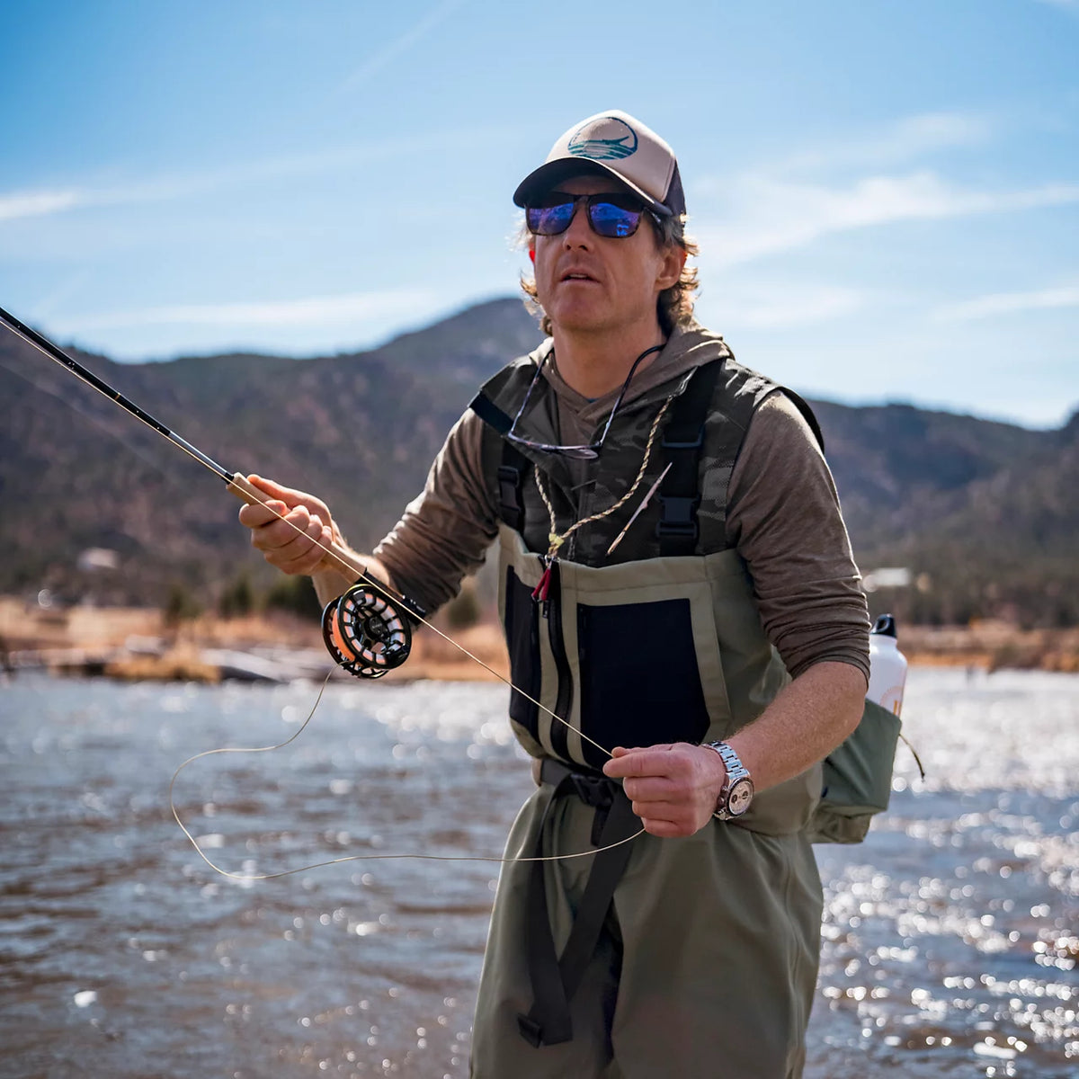 Man wearing tan Orvis Clearwater stockingfoot waders fly fishing in river with mountains in background