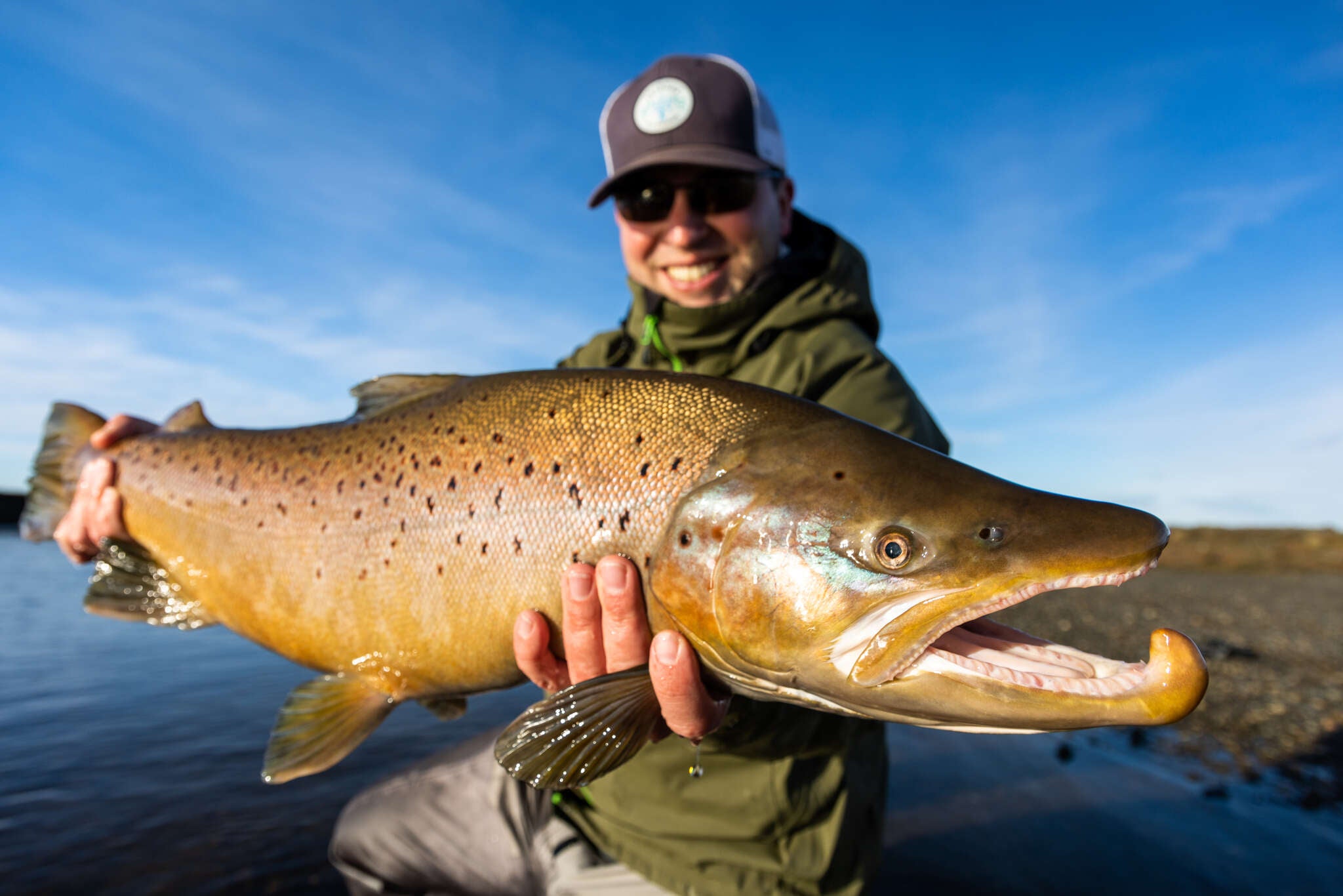 Angler in olive jacket and cap holding a large golden-brown speckled trout over a blue river, fishing