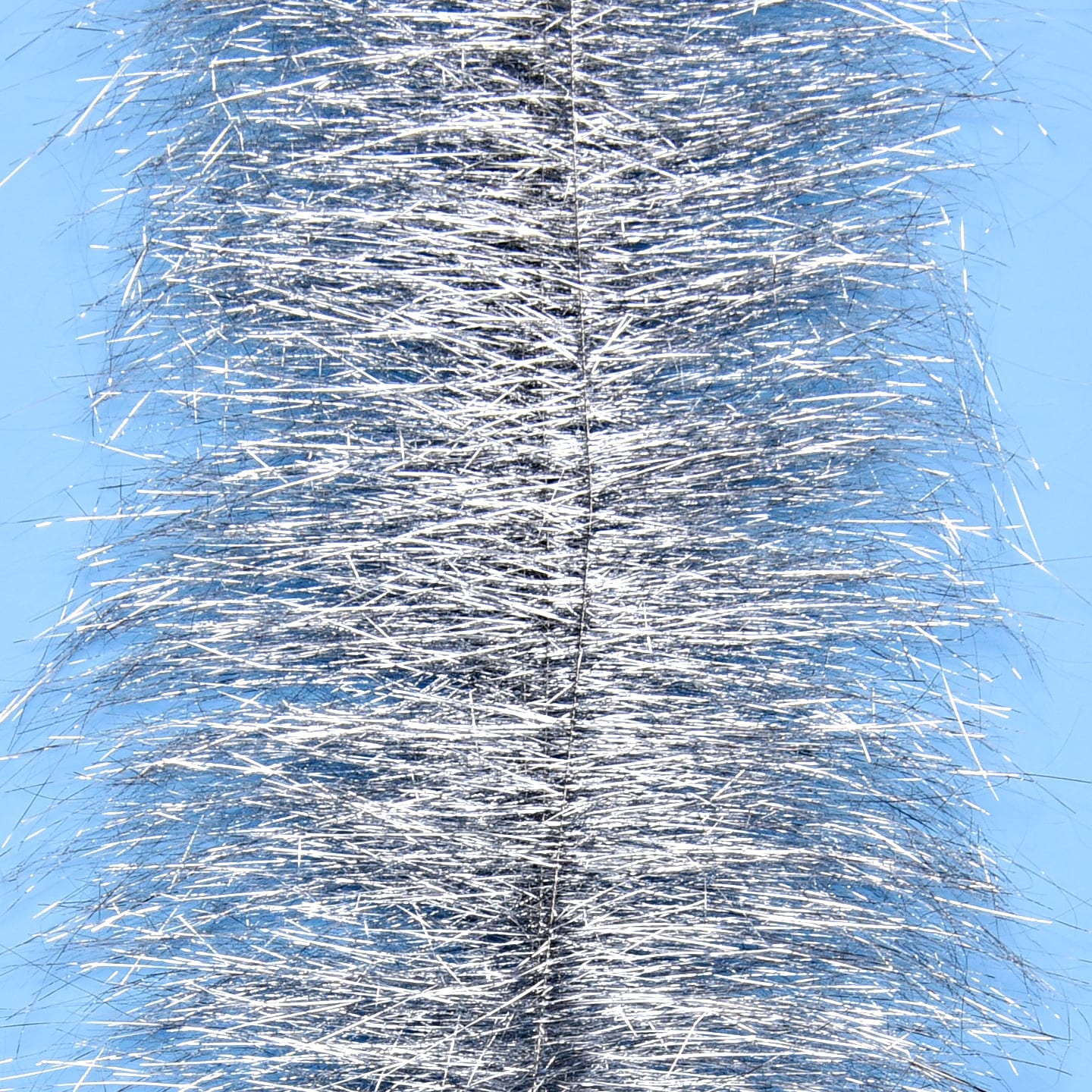 Silver metallic tinsel on dense sparkle brush for fly tying, shimmering strands against pale blue background