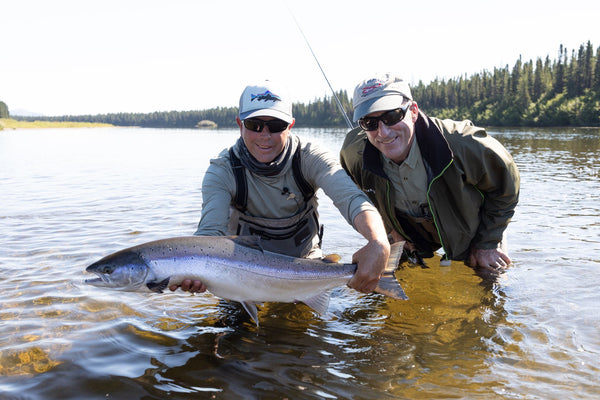 Two anglers holding large Atlantic salmon in shallow river water at Flowers River Lodge with boreal forest backdrop