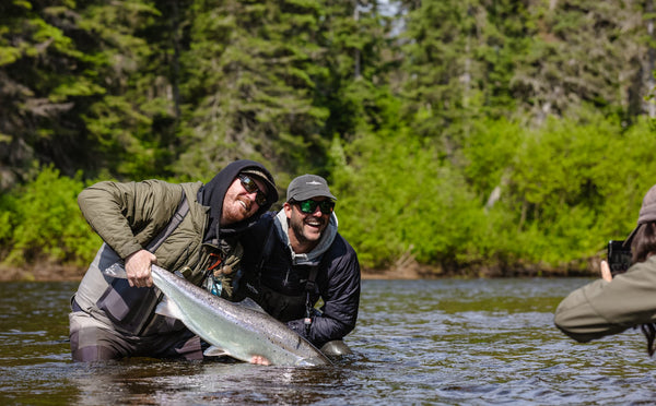 Two anglers in olive quilted jacket and dark puffer waders holding a large silver salmon on a coastal fishing trip