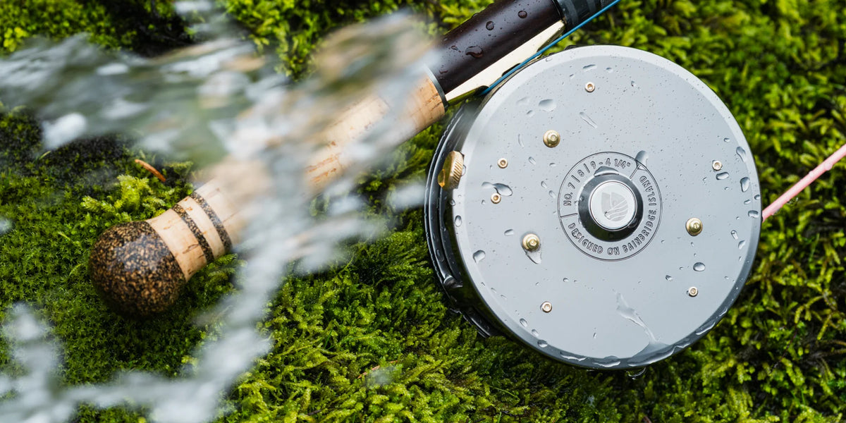 Redington Spey fly reel gray with brass hardware and water droplets on green moss beside cork rod handle