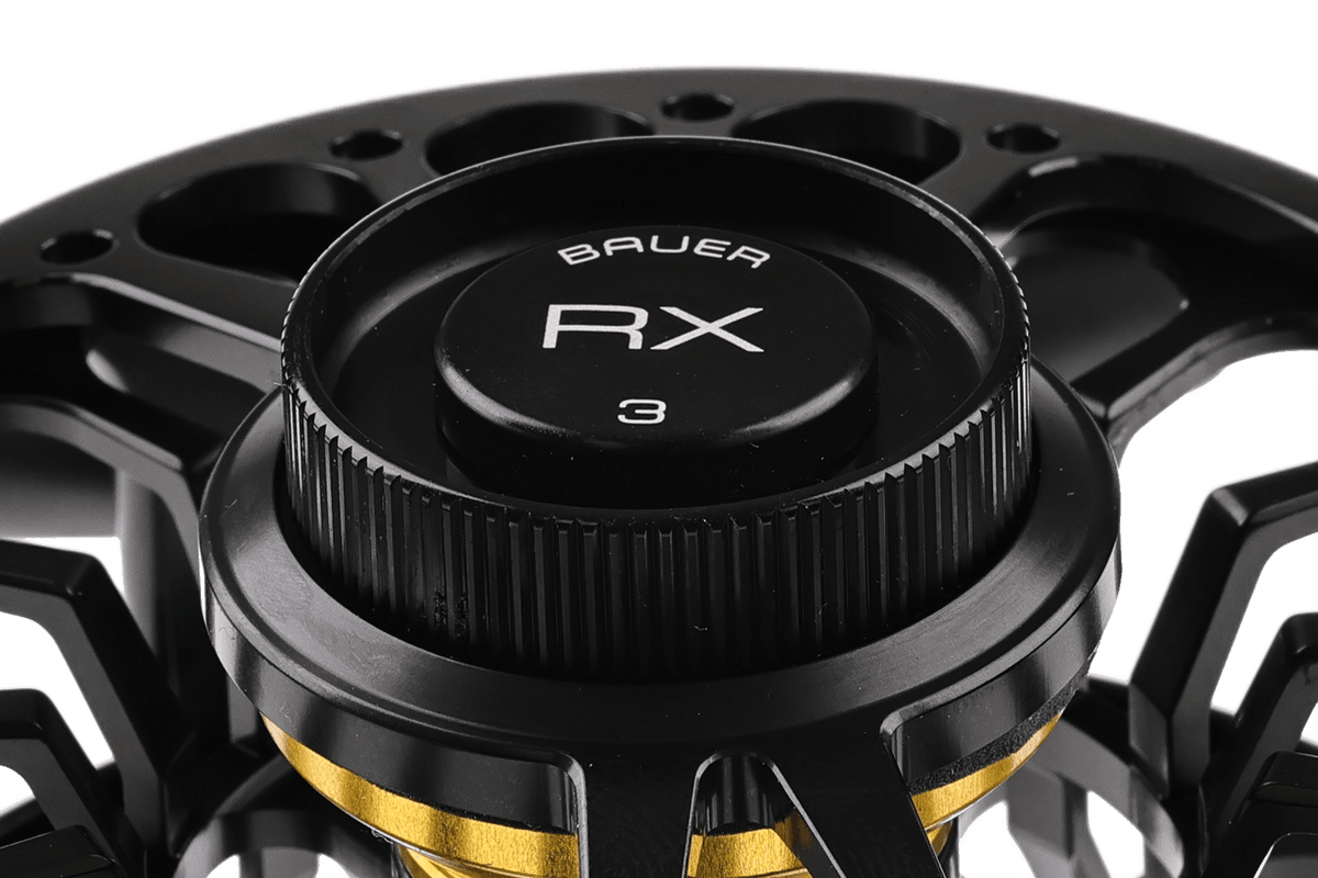 Bauer RX fly reel close-up showing black anodized machined spool, knurled drag knob labeled RX and gold accent ring